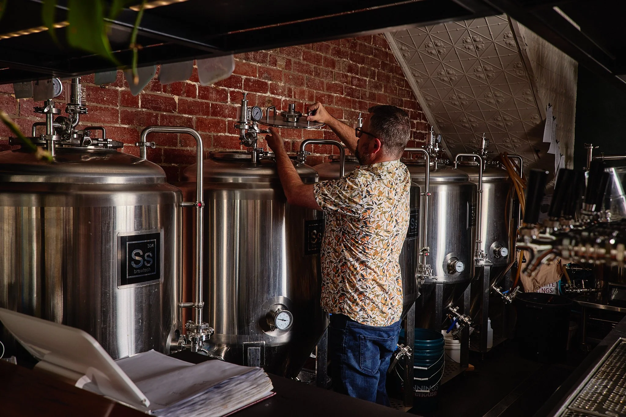 A man in a floral shirt working with large stainless steel brewing tanks in a brewery. The tanks have gauges and pipes attached, and the wall behind is made of brick.