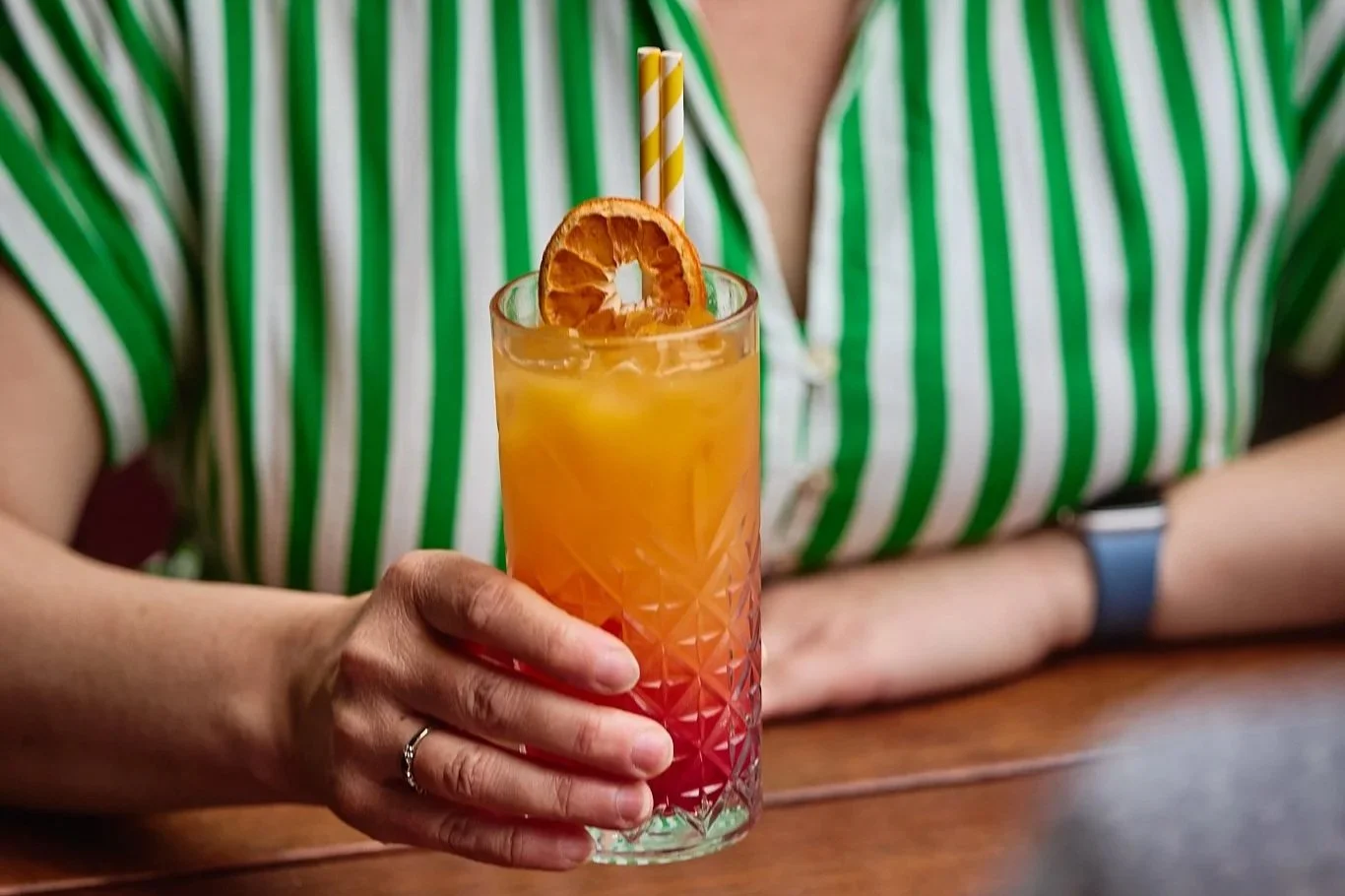 Person holding a tall glass of orange-pink layered cocktail garnished with a slice of orange and two paper straws with yellow and white stripes, sitting at a wooden table.
