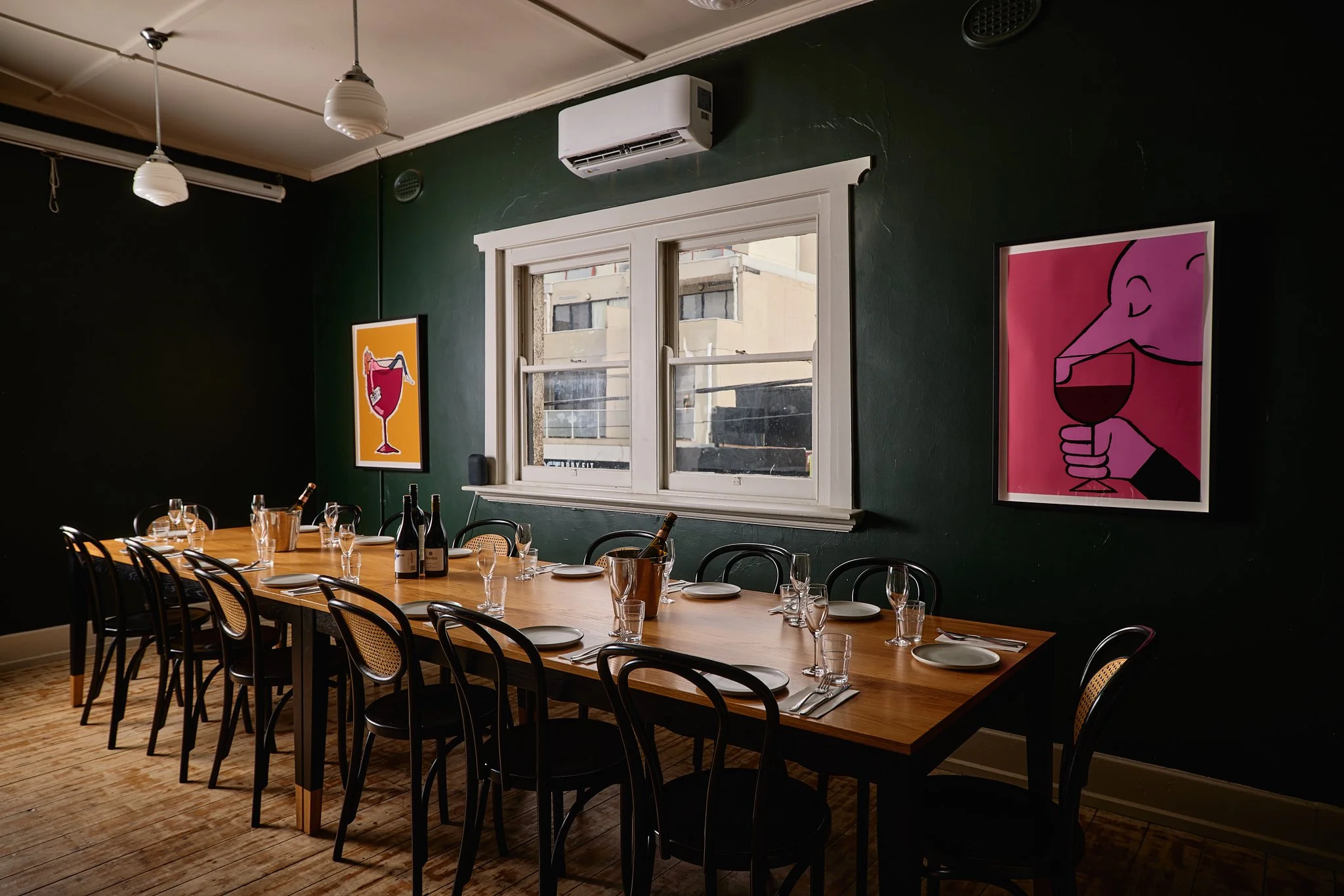 A dining room with a long wooden table set with plates, glasses, and wine bottles. There are black chairs around the table. The room has dark green walls with colorful framed art and a window above the table.