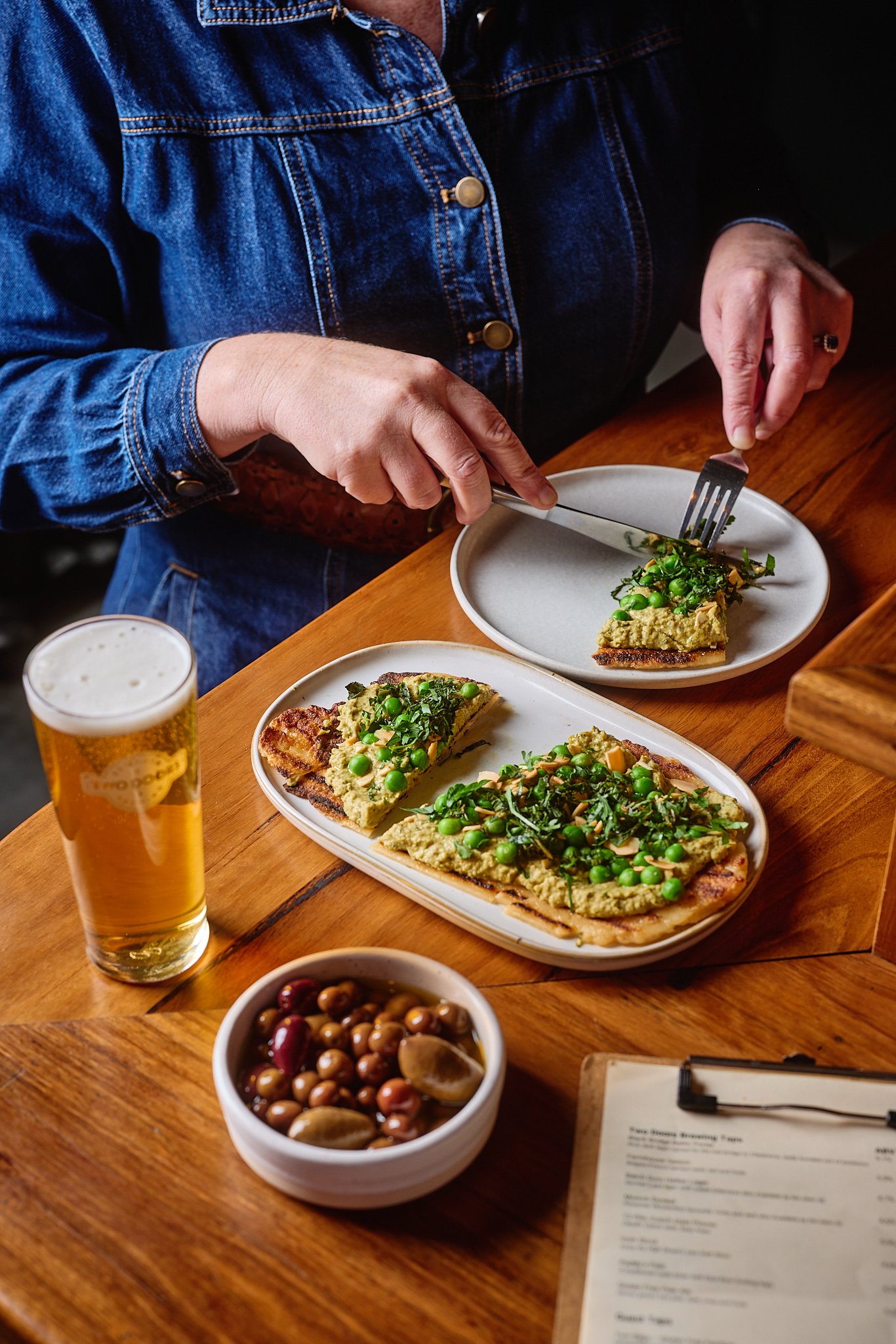 Person in denim jacket cutting avocado toast topped with peas and herbs, with a glass of beer and a bowl of mixed olives on a wooden table.