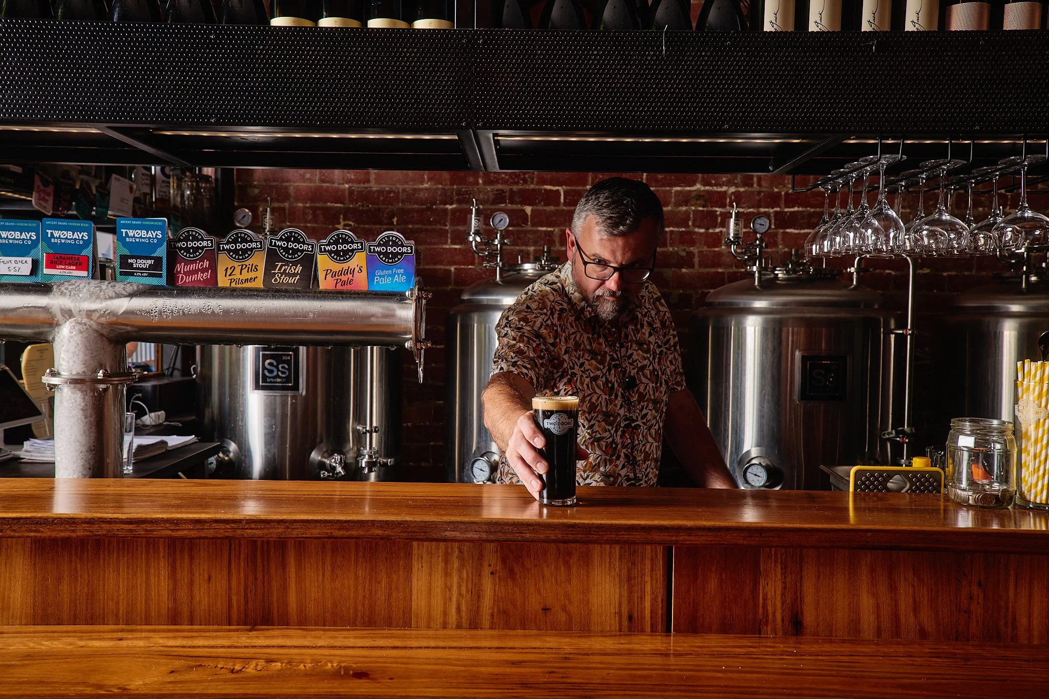 A bartender with glasses and a patterned shirt pouring a dark beer at a brewery bar with brewery tanks and brick wall background.