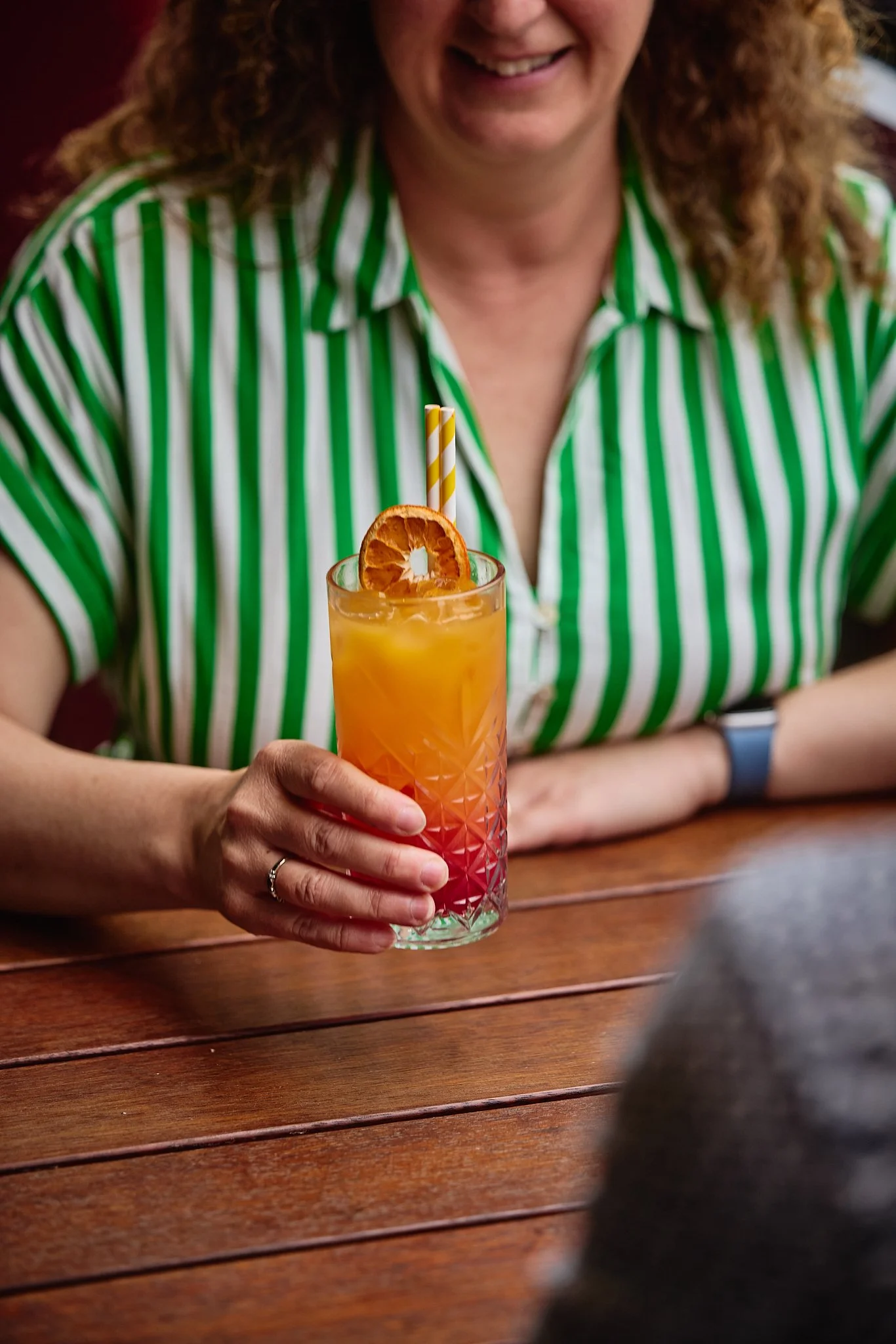 Woman in a green and white striped shirt holding a colorful cocktail with an orange slice and a striped straw, sitting at a wooden table.