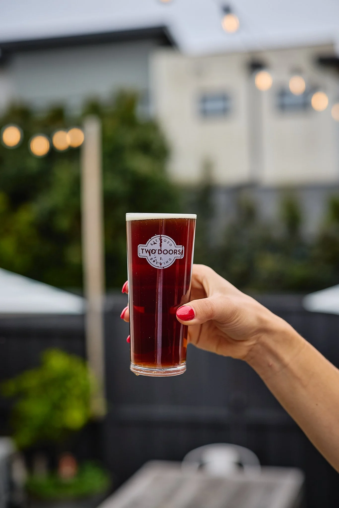 A person with red painted nails holding a tall glass of dark beer with a white logo that says 'Two Doors Brewing' in an outdoor setting with bokeh string lights and a house in the background.