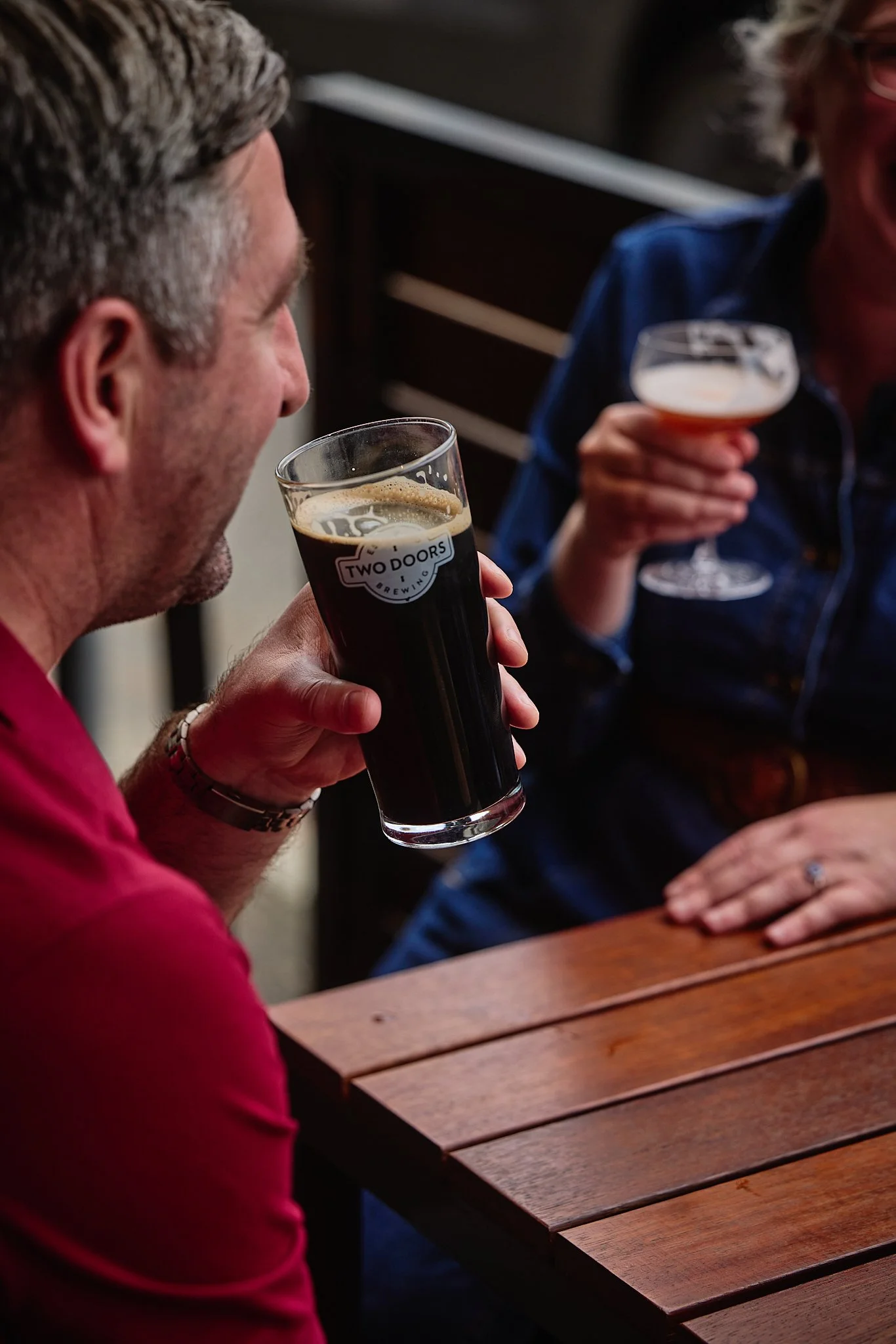 Two people sitting at a wooden table, each holding a different glass of beer, engaging in a conversation. One is wearing a red shirt, and the other is wearing a blue jacket.