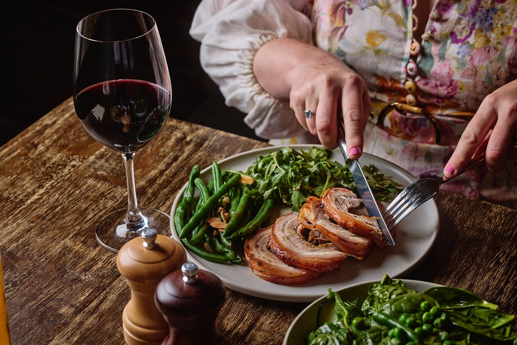 A person is slicing cooked pork belly on a plate filled with green beans, salad, and other vegetables, with a glass of red wine and salt and pepper shakers on a rustic wooden table.