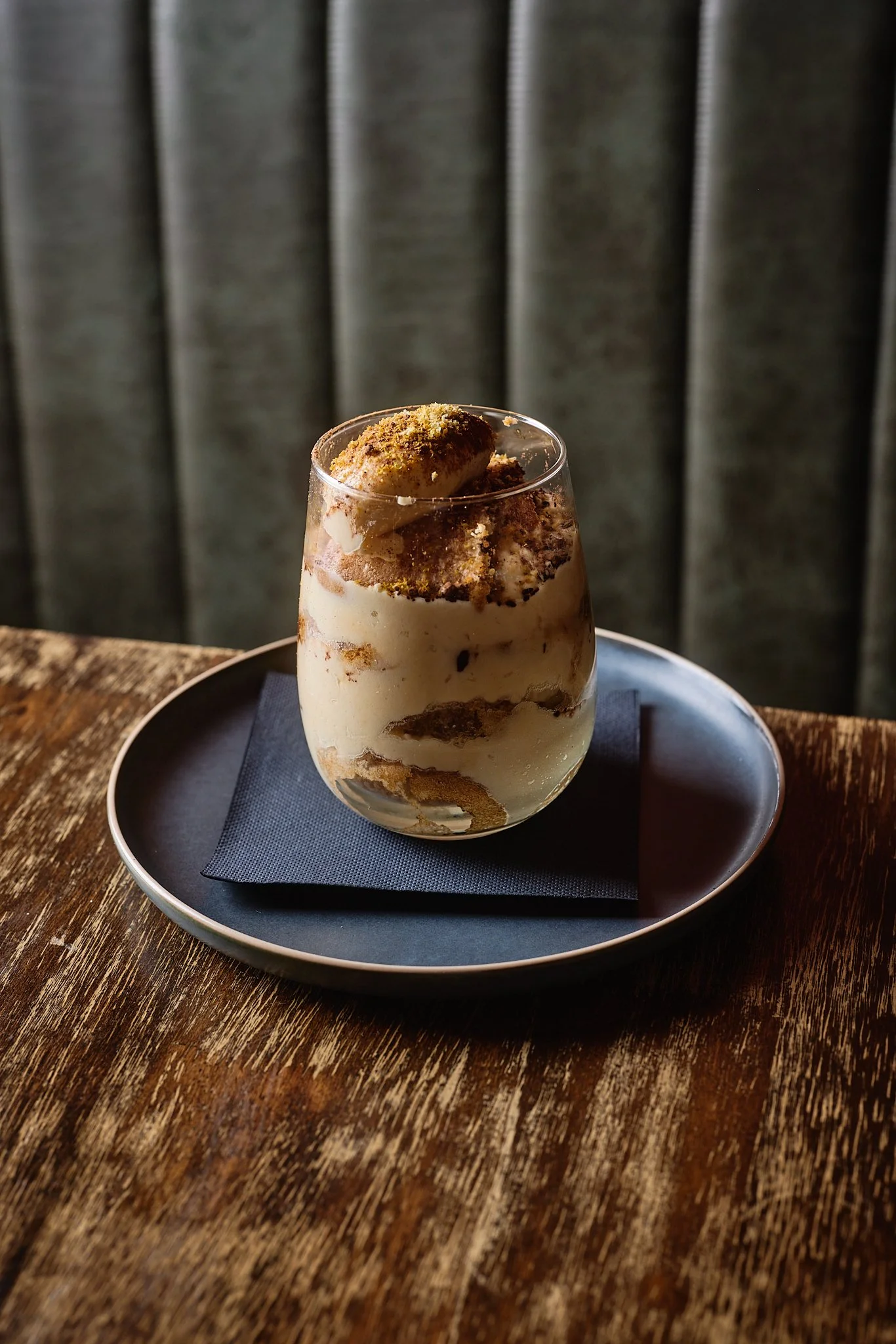 A glass of tiramisu layered with cream, coffee-soaked ladyfingers, and cocoa powder on top, served on a dark plate with a napkin, on a wooden table.