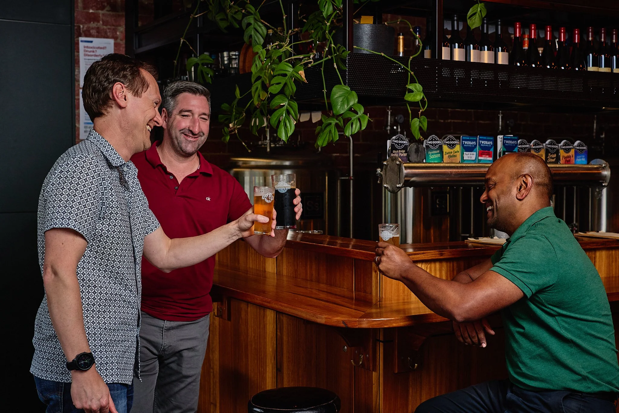Three men in a bar smiling, two are standing and one is sitting, holding glasses of beer for a toast.