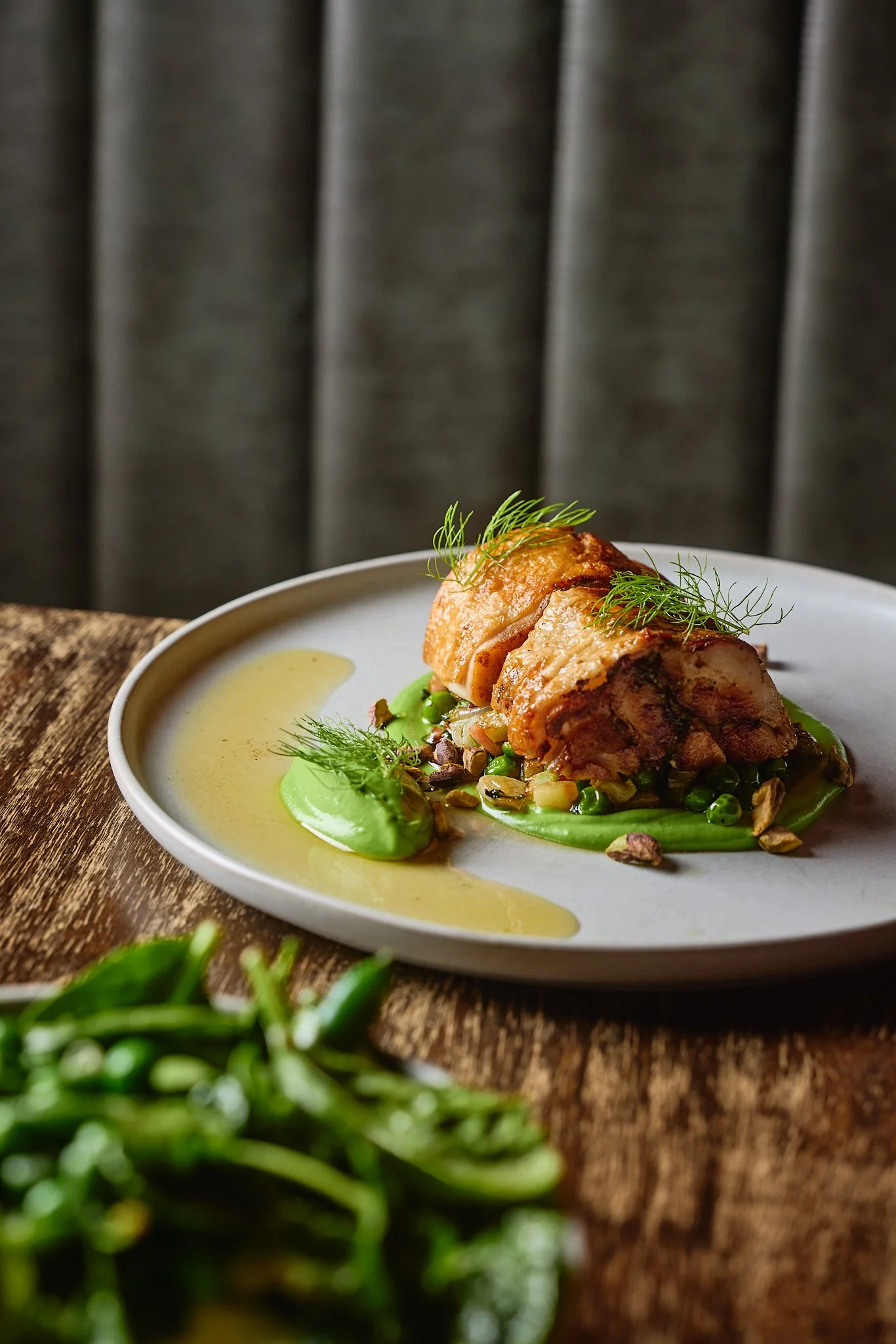 A plated dish featuring crispy roasted chicken, green pea puree, garnished with herbs and seeds, with a sauce on the side, on a rustic wooden table.