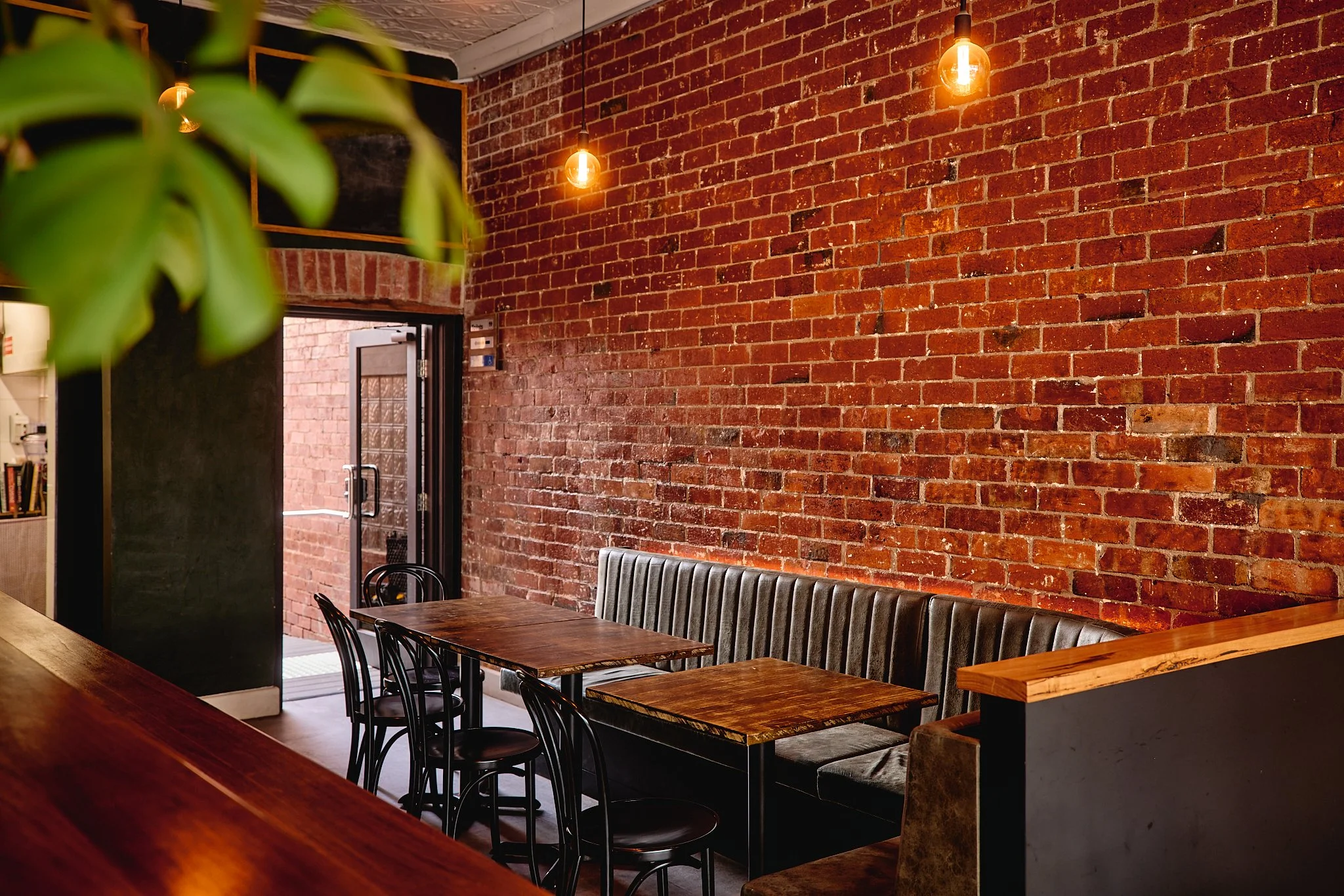 Interior of a trendy cafe with an exposed brick wall, hanging Edison-style light bulbs, a wooden table, black chairs, and a black leather bench seating. A door leads to an outdoor area.