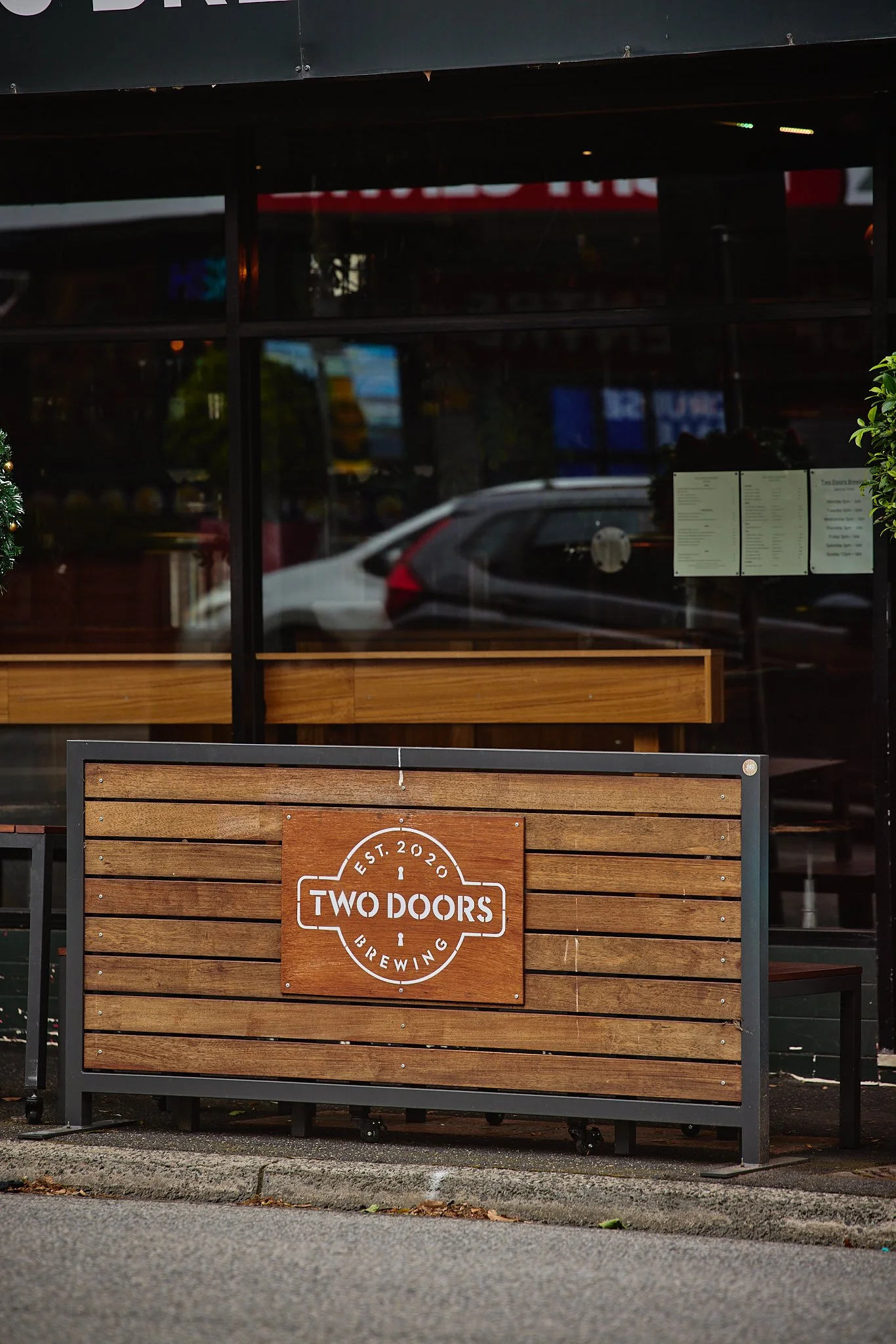 Wooden sign with the logo of Two Doors Brewing in front of a glass storefront with cars reflected in the window.
