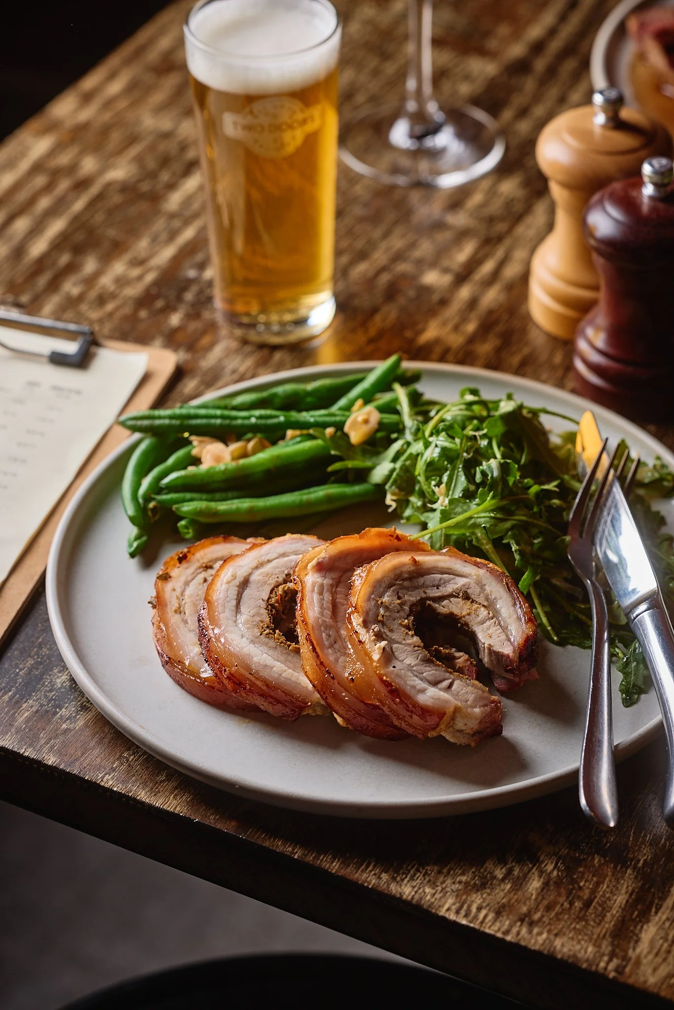 A plate of roasted pork belly with green beans, arugula salad, and garlic, accompanied by a glass of beer on a rustic wooden table.