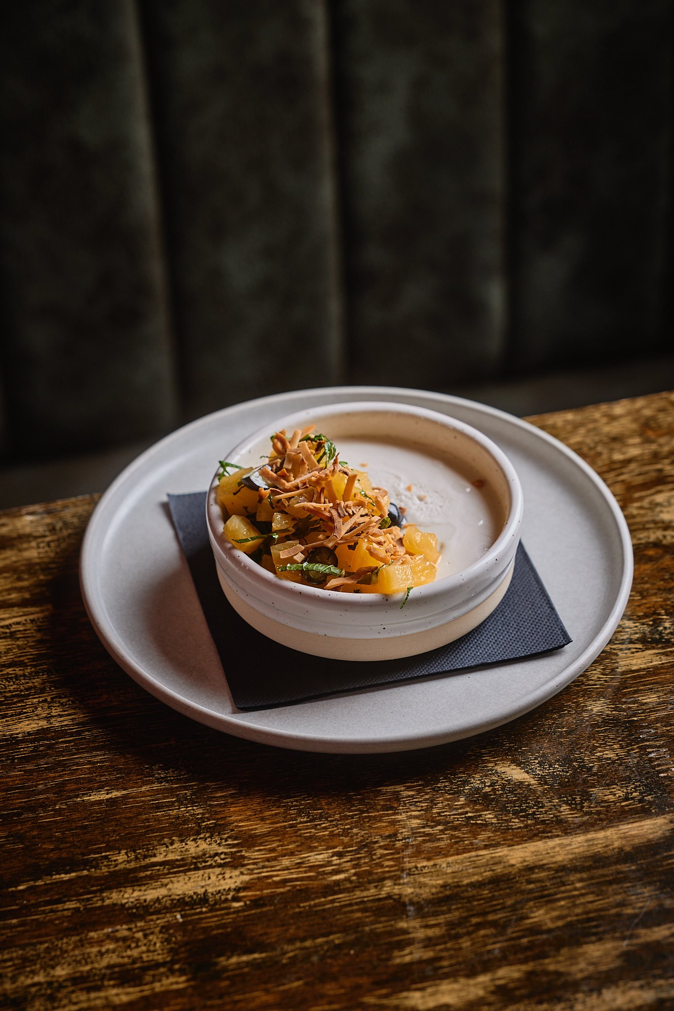 A bowl of creamy yogurt topped with diced pineapple, chopped nuts, and herbs, served on a dark napkin on a light plate, placed on a rustic wooden table.