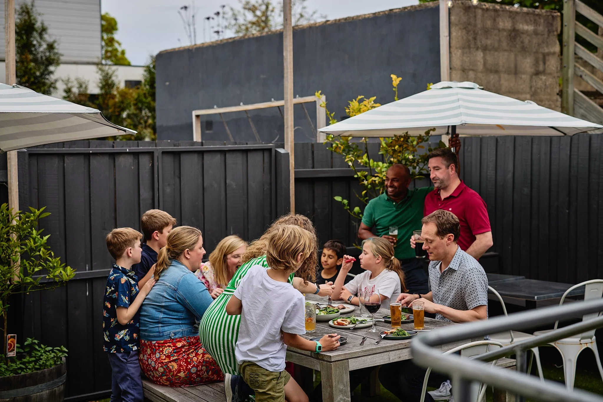 People having a backyard gathering around a table with food and drinks, two men stand by umbrellas, kids and adults sit and chat near a black fence.