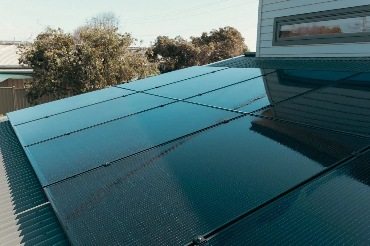 A row of installed solar panels on a house roof during daytime, with trees and a house in the background.