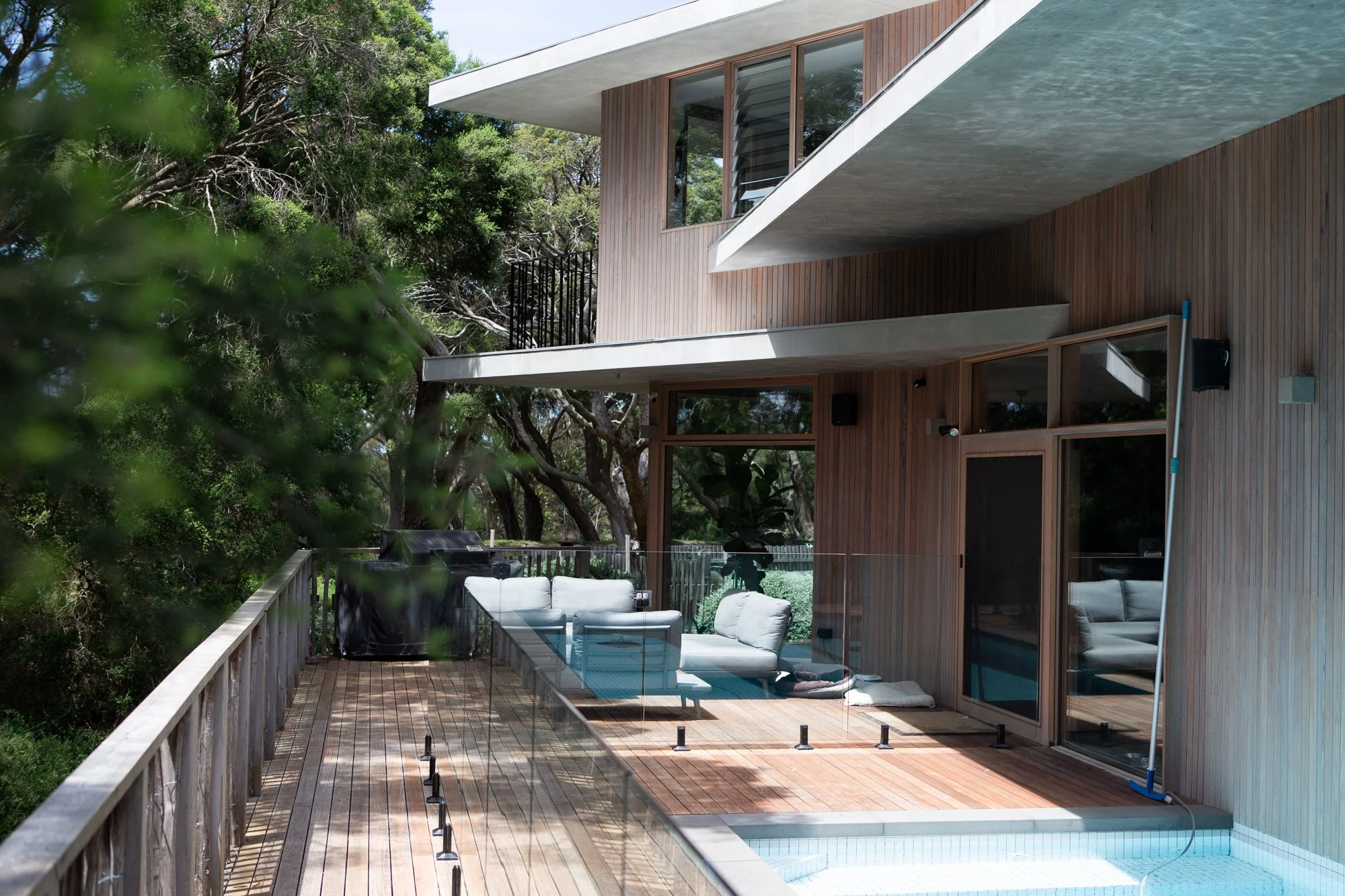 Modern house with wooden exterior, sliding glass doors, outdoor sitting area with white couches, glass railing, and a swimming pool with a pool cleaning pole, surrounded by trees.
