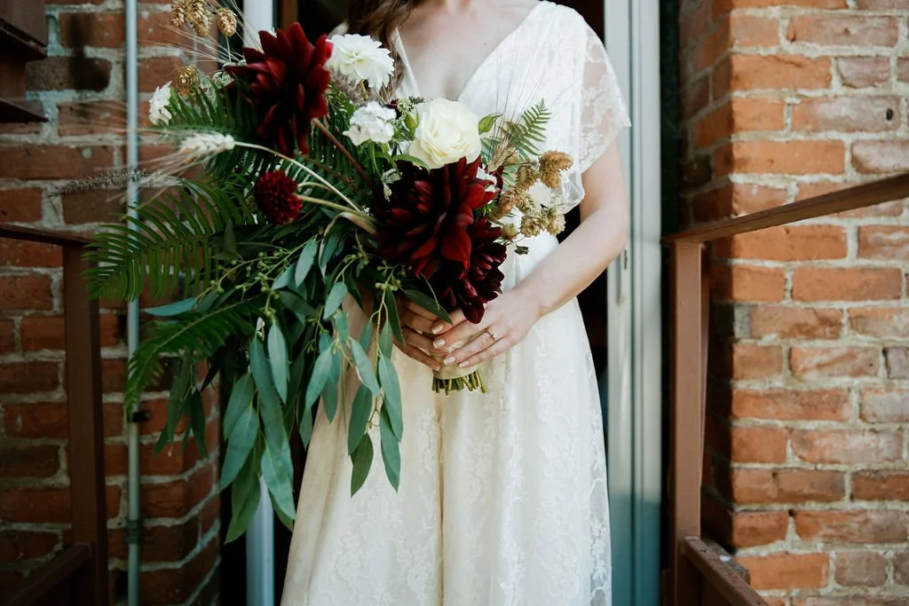 A woman in a white lace dress holding a large bouquet of flowers that includes red dahlias, white roses, and green foliage, standing on a small balcony with brick walls.