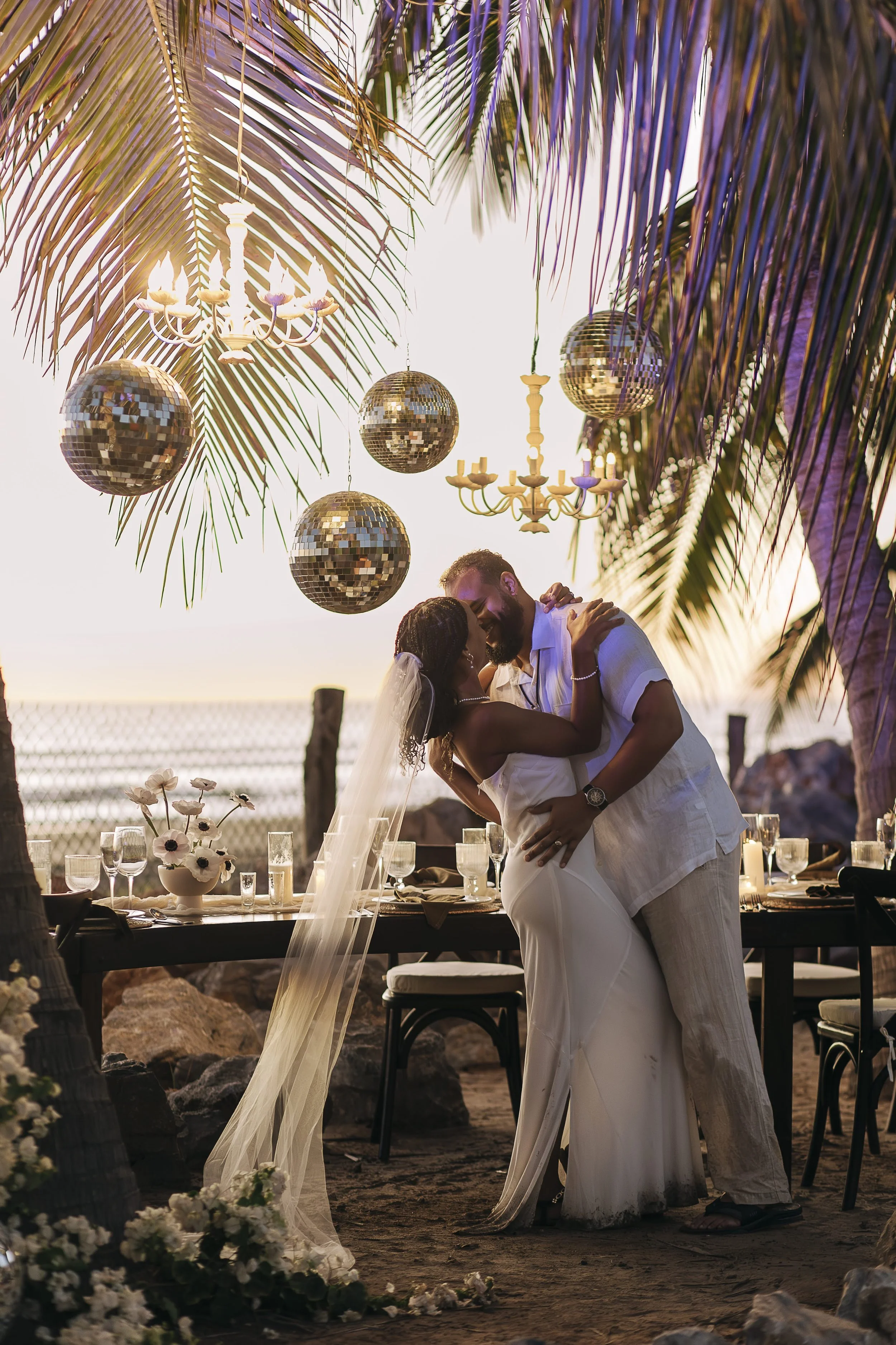 A couple sharing a dance at a beach wedding reception during sunset, with palm trees, disco balls, chandeliers, and a decorated dining table in the background.
