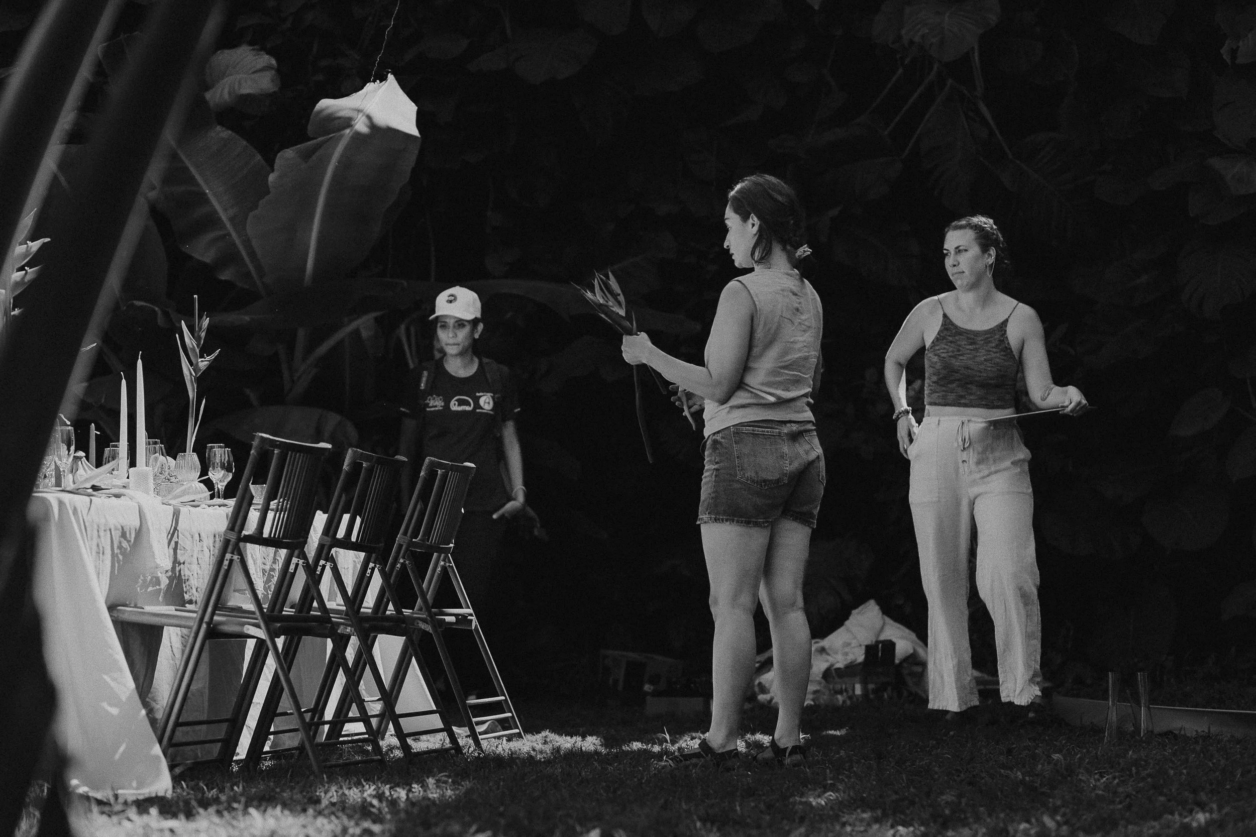 Two women and Sarah preparing a dinner table outdoors in a garden, with large leafy plants in the background, in black and white.