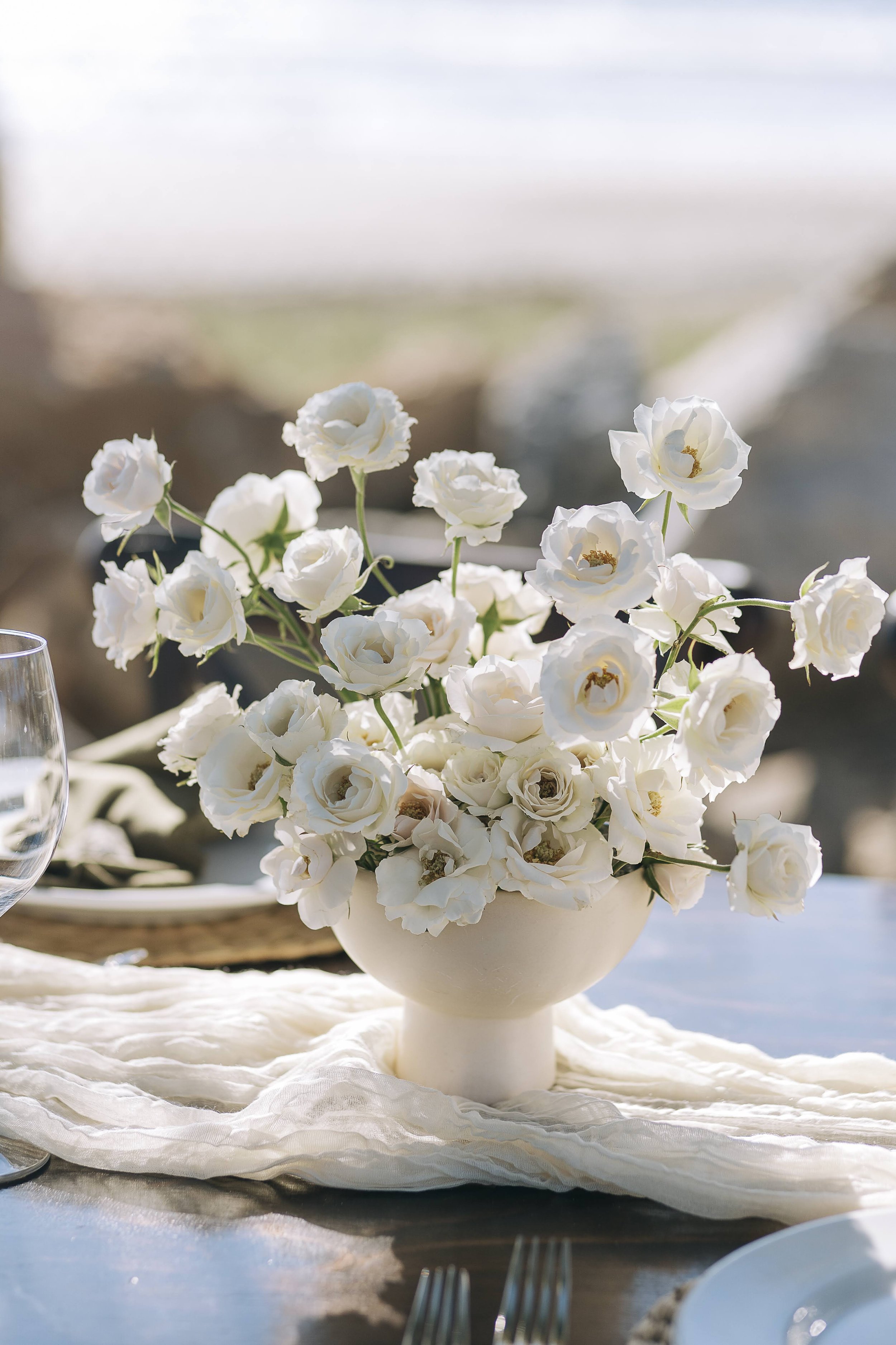A white vase filled with white flowers, placed on a table with a white cloth, set for a meal outdoors.