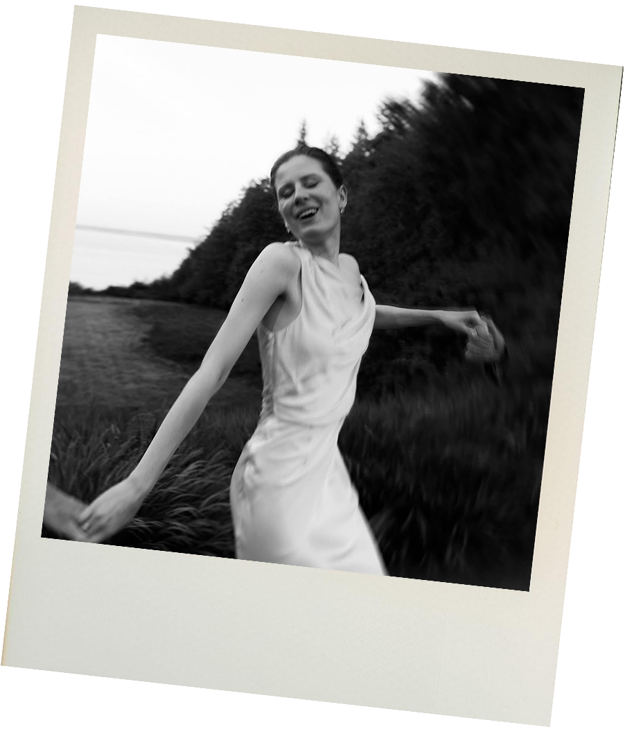 A woman in a light-colored dress smiling and enjoying herself outdoors in a black-and-white photo, with a background of trees and a grassy area.