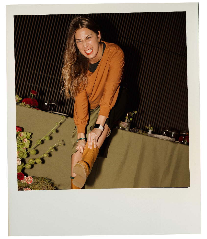 Sarah with long wavy hair wearing an orange top and beige boots, smiling and squinting while holding her booted leg up, in front of a table decorated with flowers and plants, with a dark patterned background.