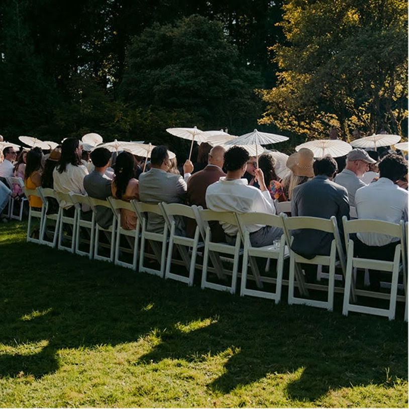 People seated outdoors at a formal event, holding white parasols, in a lush green garden in the late afternoon.