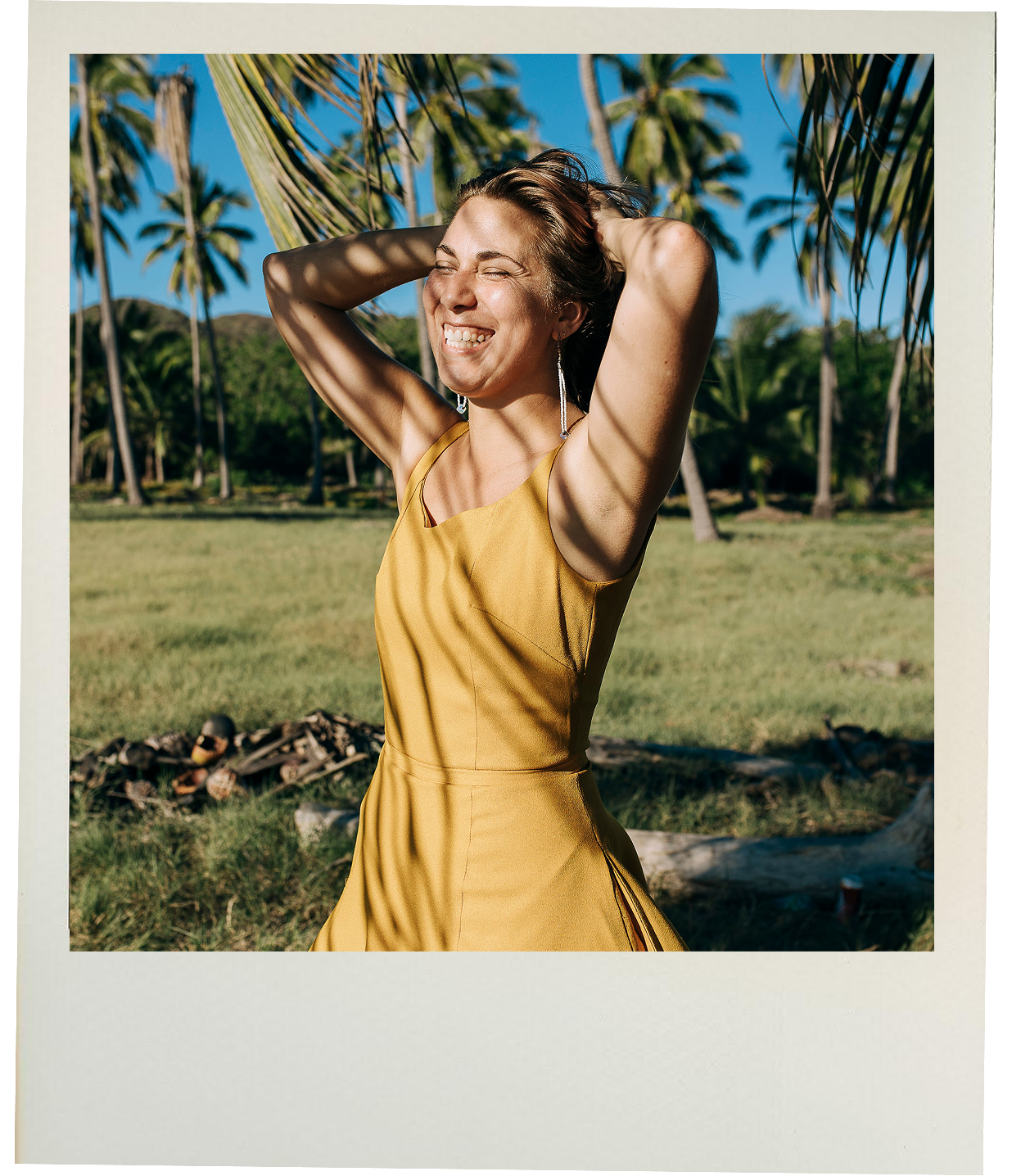 Sarah smiling with eyes closed and hands behind head, wearing a yellow dress outdoors with palm trees and green foliage in the background in Mexico.