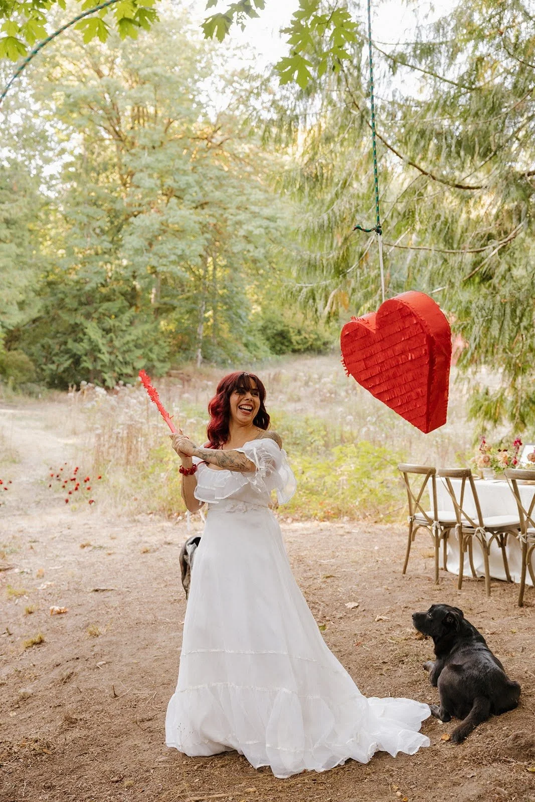 Woman wearing a white dress holding a red heart-shaped piñata stick, smiling, outdoors with a black dog sitting nearby, and a large red heart-shaped piñata hanging from a tree branch, surrounded by chairs and a table with flowers.