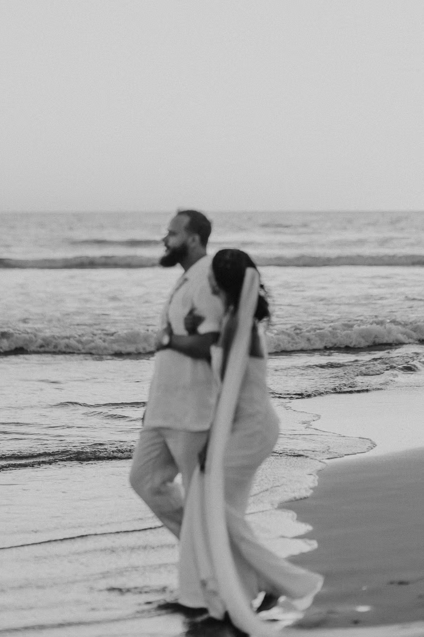 A man and a woman walk along the beach. Both are dressed in light clothing. The scene is black and white, with the ocean and waves in the background.