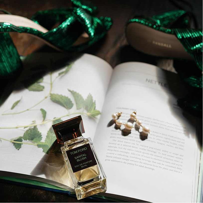 Open magazine with green leaves on the pages, a pair of pearl earrings on the right page, a small bottle of Tom Ford Santal Blush perfume on the left page, and green ribbon decorations around the magazine.