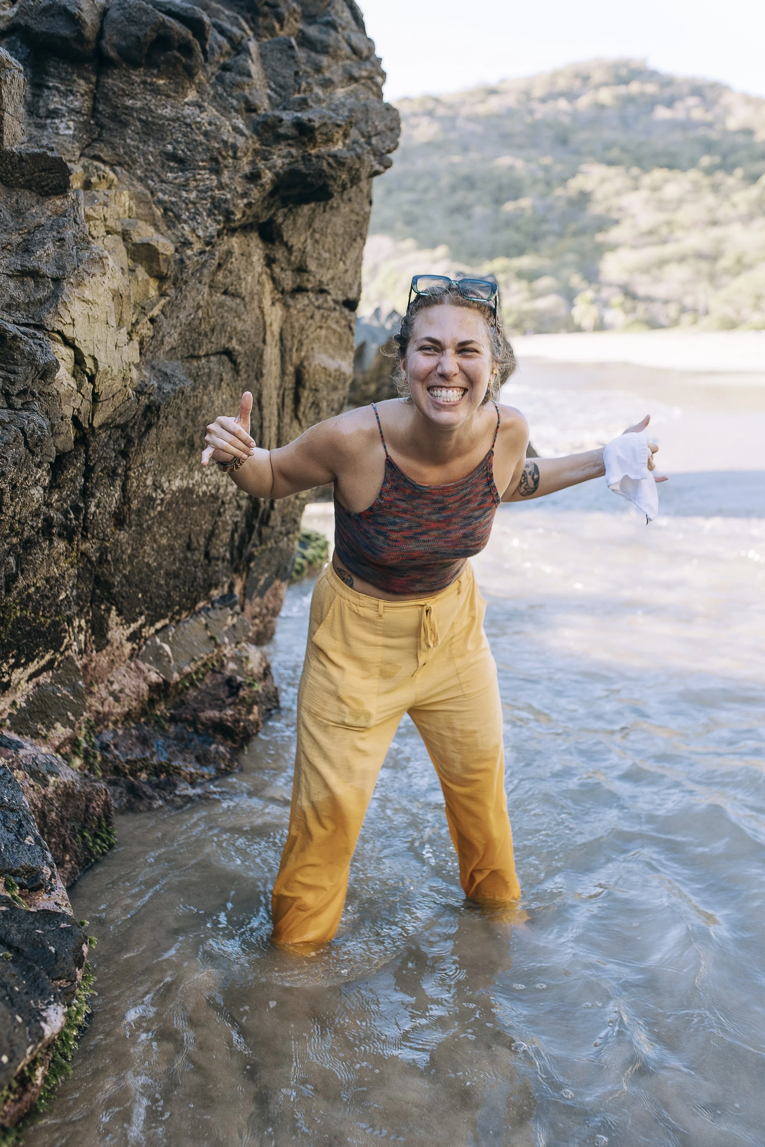 Sarah in yellow pants and a colorful tank top smiling and making a playful gesture on a rocky beach with water and a hillside in the background.