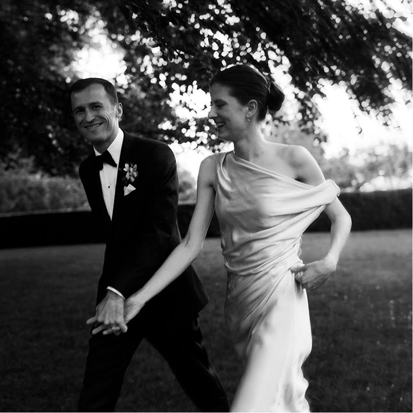 A happy couple, dressed in wedding attire, holding hands and walking outdoors under trees in black and white.