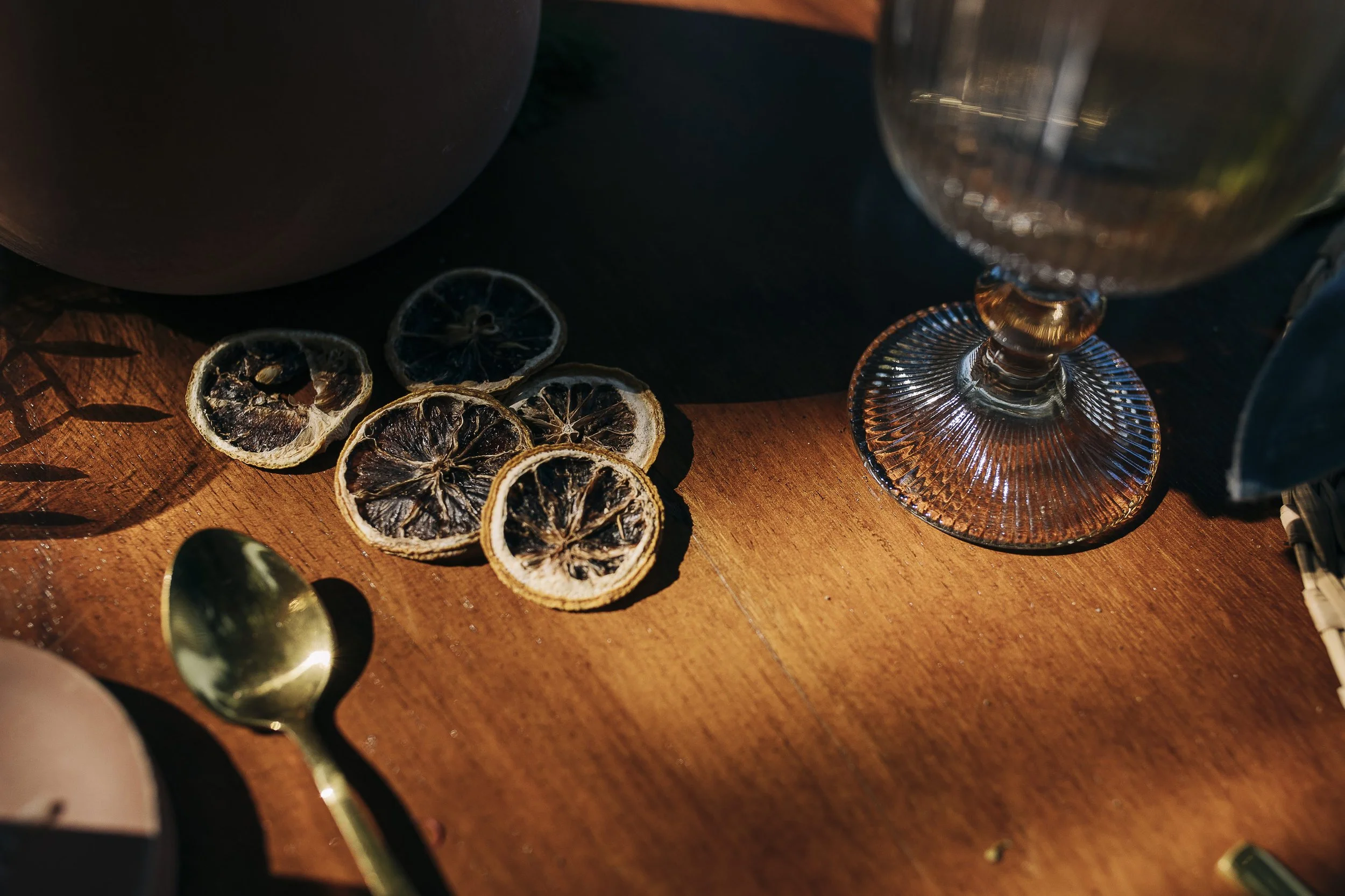 Dried lemon slices on a wooden surface next to a glass of water and a gold spoon, with shadows cast by nearby objects.