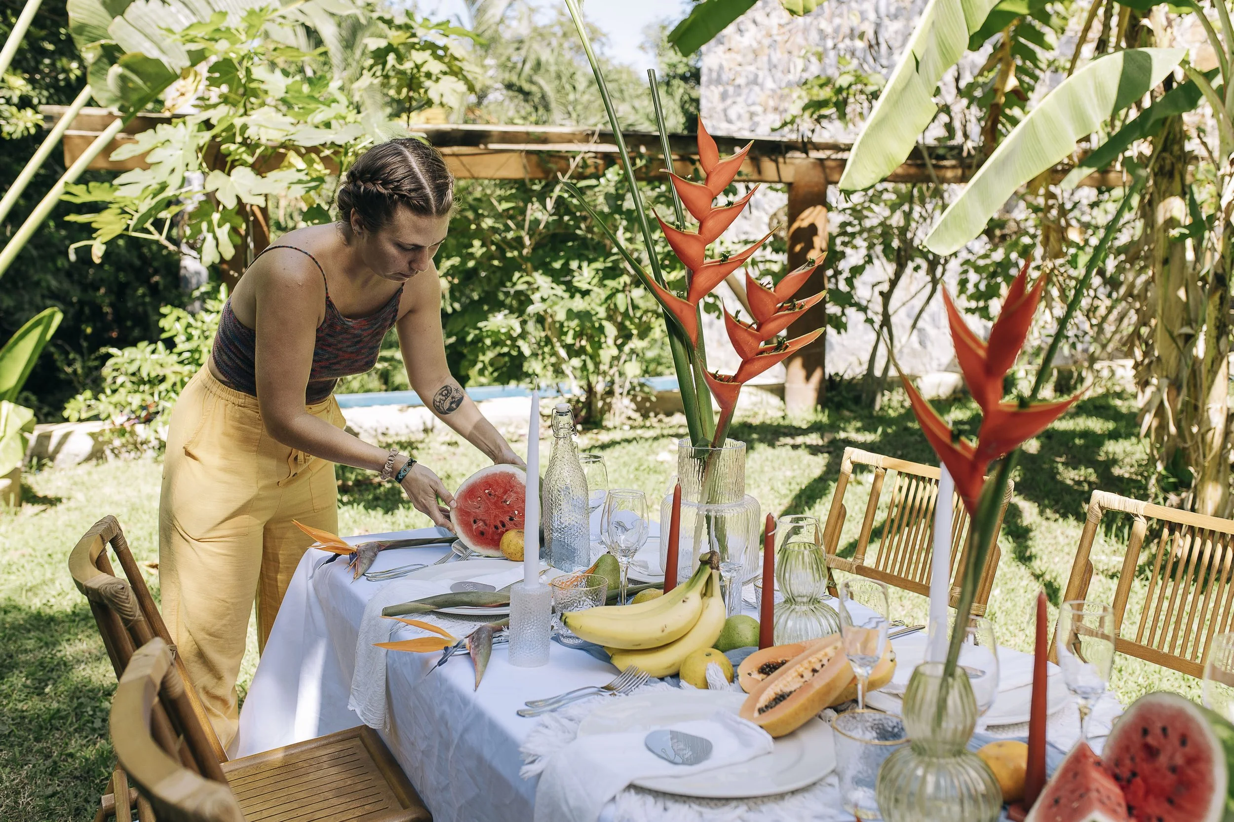 Sarah with braided hair and a tattoo on her arm setting a watermelon slice on a table decorated with tropical fruits, floral arrangements, candles, and glassware in an outdoor garden.