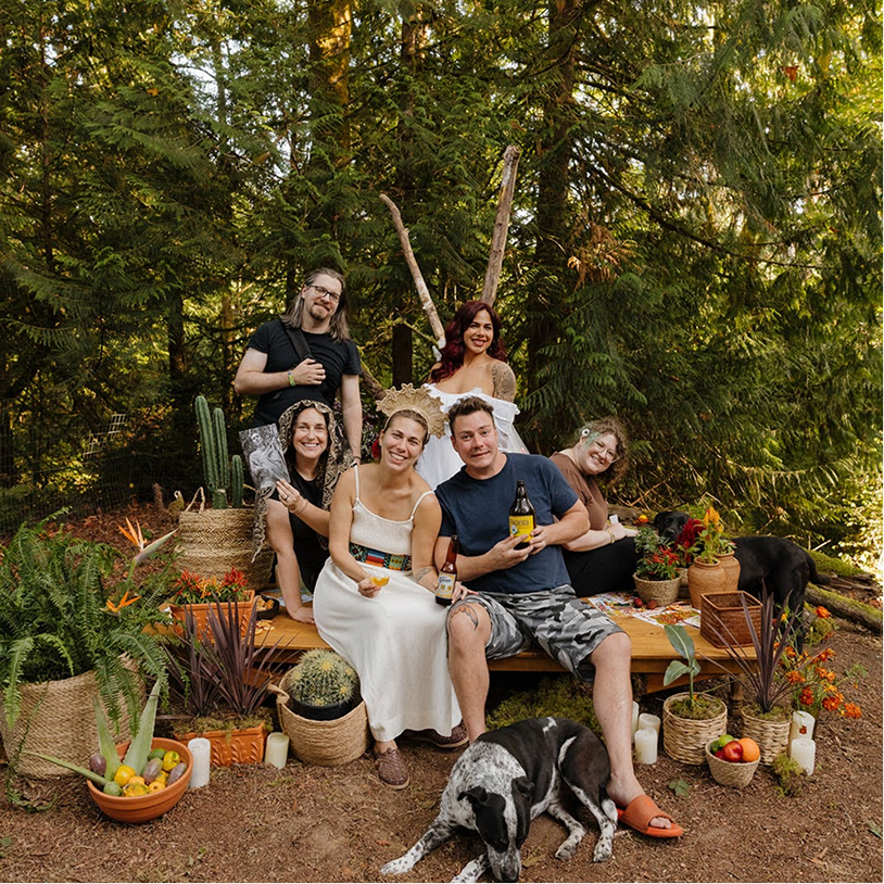 Group of six friends outdoors, sitting and standing on a wooden platform surrounded by plants and candles, with a decorative tree and forest background, friends smiling, some holding drinks, dogs lying on the ground.