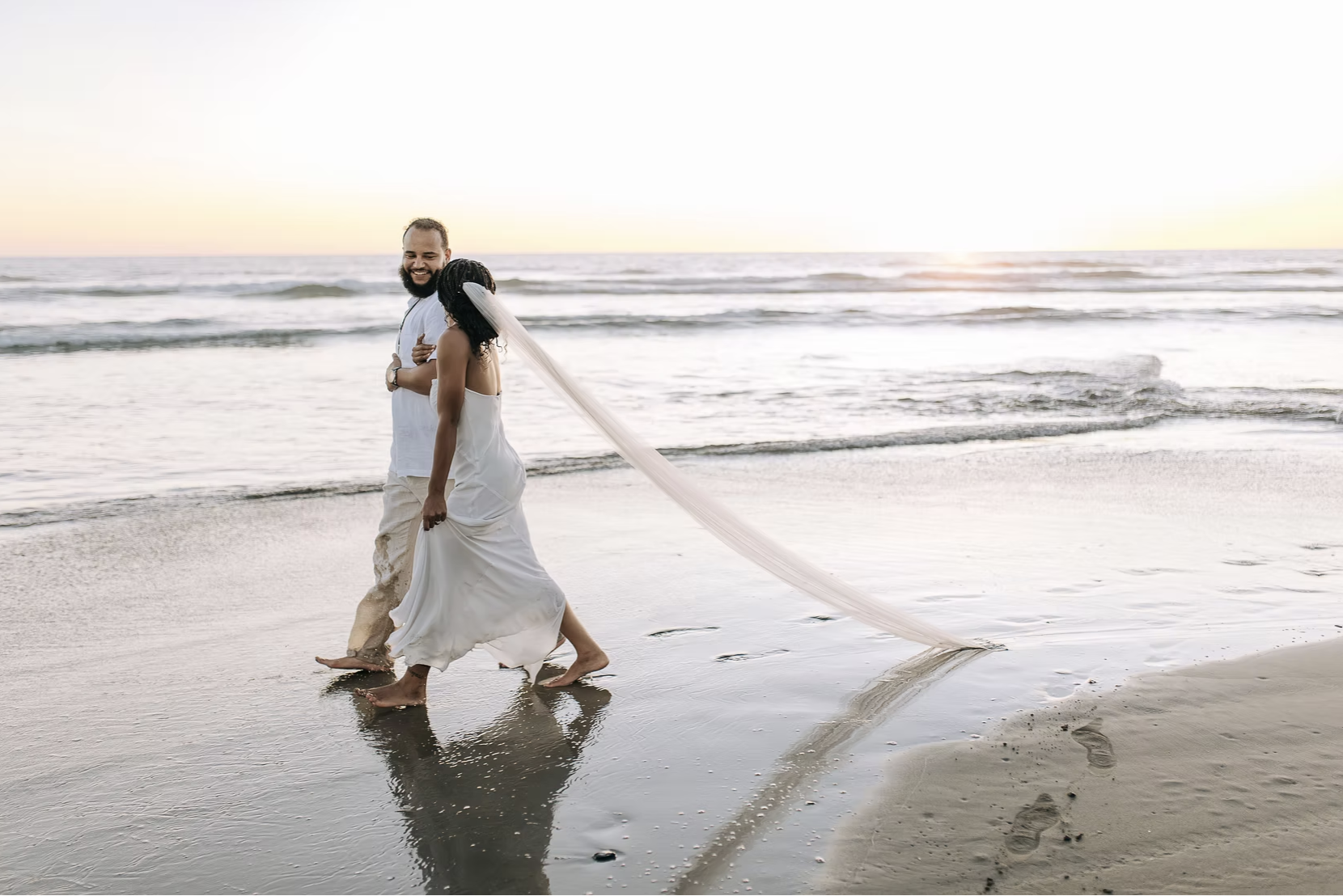 A newlywed couple walking barefoot on the beach at sunset, with the bride in a white dress and veil, and the groom in a white shirt and beige pants, holding hands and smiling.