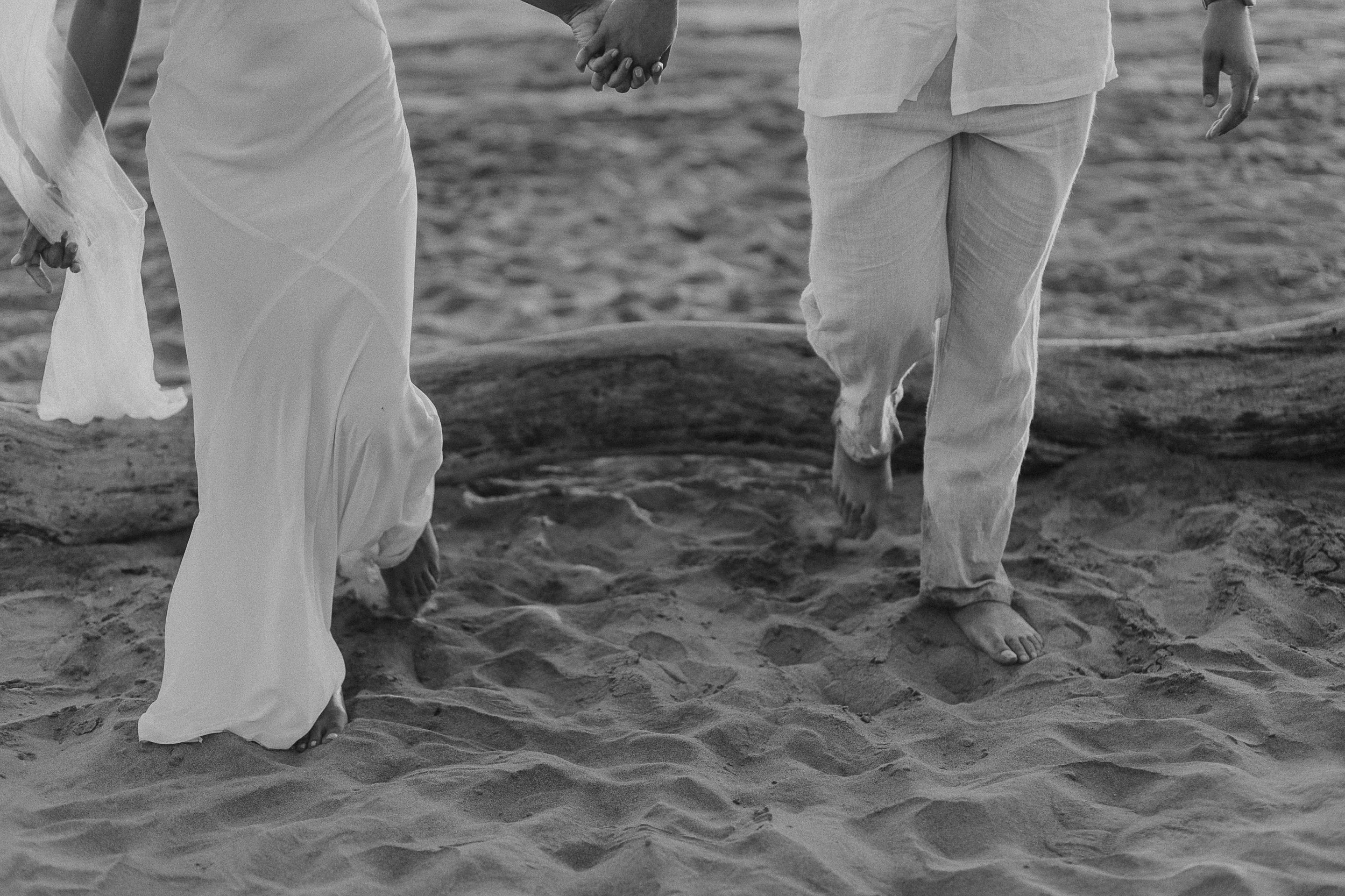 Close-up of a couple holding hands as they walk barefoot on the sandy beach near the water, with one wearing a long white dress and the other in light-colored pants and shirt.