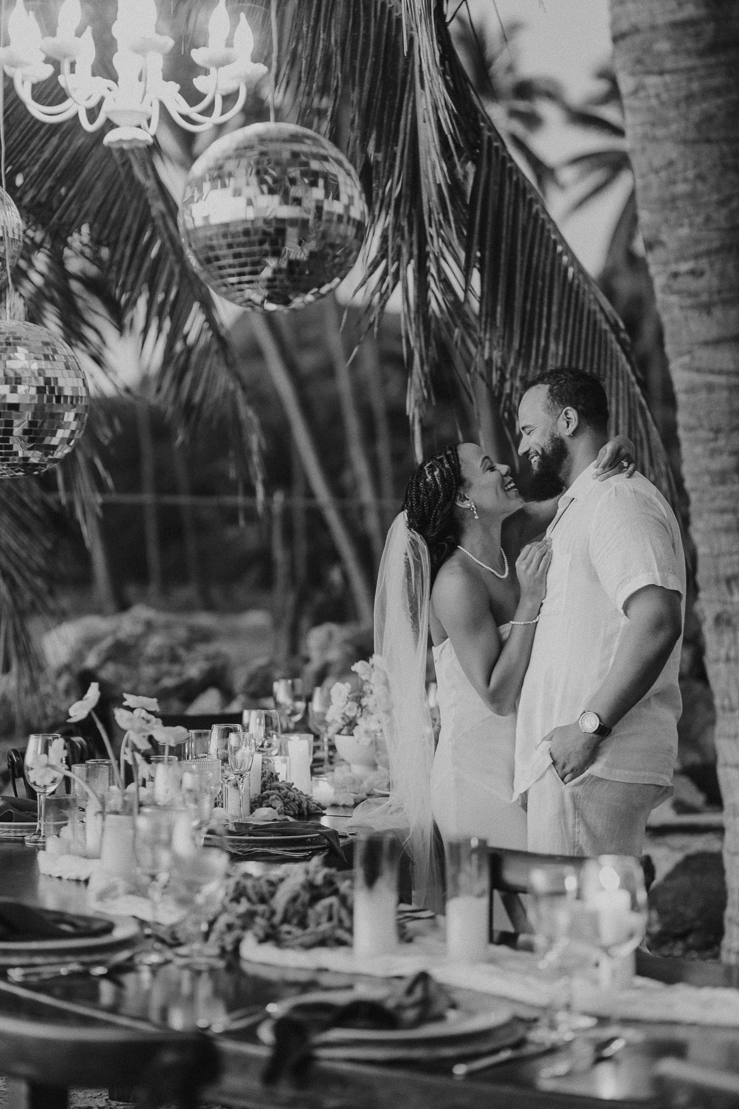 Black and white photo of a couples' wedding reception, with the bride and groom smiling and embracing under hanging disco balls and decorations, surrounded by a beautifully set table with candles and flowers.