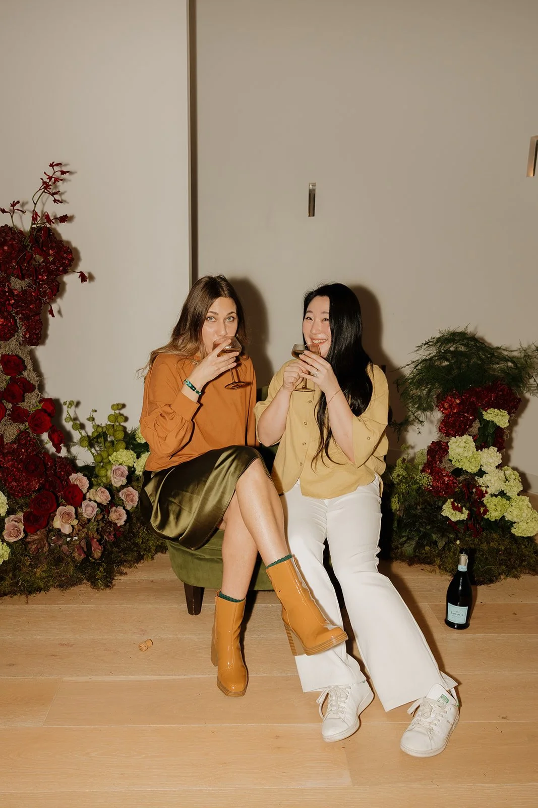 Two women sitting on a green bench, sharing drinks and smiling. One wears a brown top and a skirt, the other wears a yellow shirt and white pants. They are surrounded by floral arrangements and a bottle of wine on the floor.
