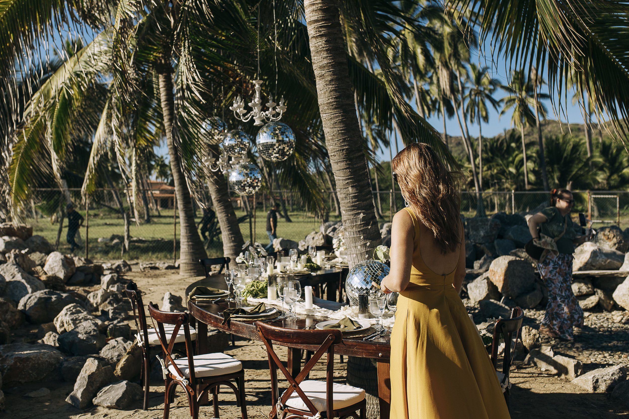 Sarah in a yellow dress standing next to a dining table set up on a beach surrounded by palm trees, with disco balls hanging from the trees and people in the background.