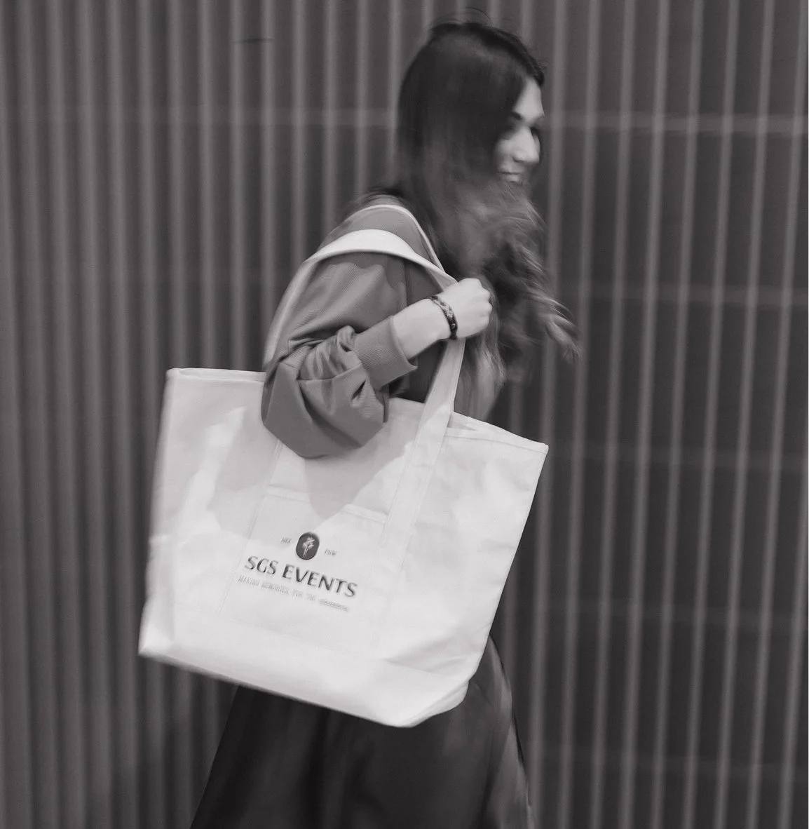 Sarah walking and carrying a large tote bag with 'SGS EVENTS' written on it, against a wooden-paneled background, in black and white.