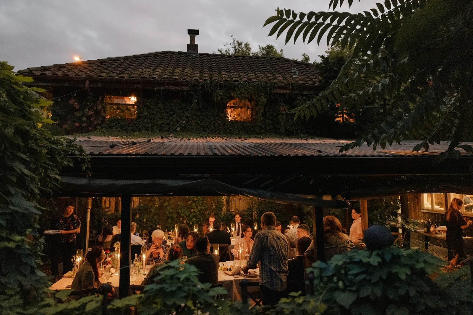 An outdoor dinner party at The Corson Building in Seattle. At dusk under a canopy, with people seated at tables decorated with candles and flowers, surrounded by lush greenery and trees, with a house in the background.
