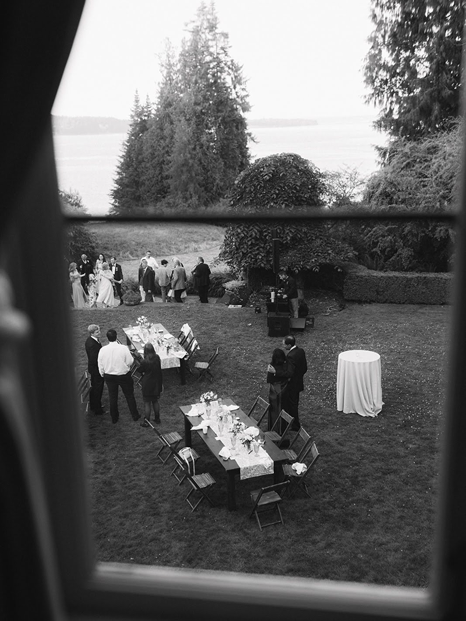 People at an outdoor party through a window at dusk, with tables and a sound system set up on a grassy area and trees in the background.