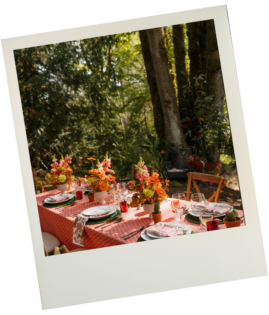 Outdoor dining table set with floral arrangements, plates, glasses, and napkins in a garden with trees and sunlight
