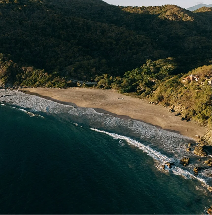 Aerial view of a beach in Mexico surrounded by green hills and dense trees, with the ocean on one side and hillside houses on the other.