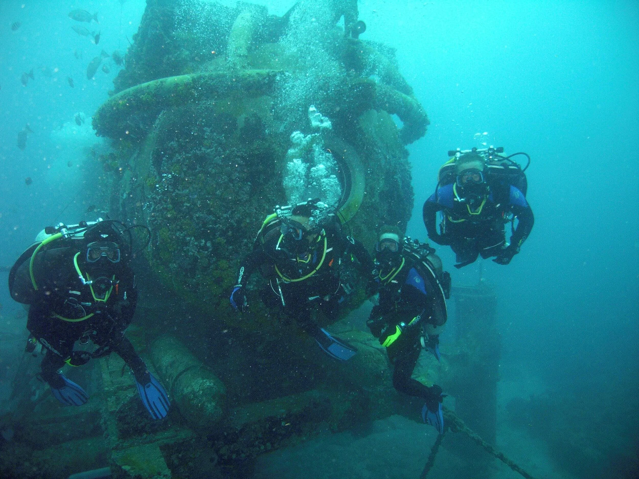 four divers in front of the NEEMO undersea habitat. some fish can be seen off the left side. The habitat already has a lot of algae growing on it