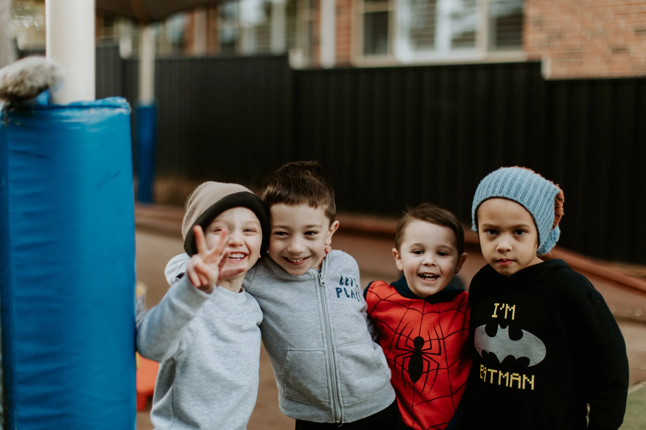Four young boys standing outside near a black fence and a brick building, smiling and posing for a photo on a cloudy day.
