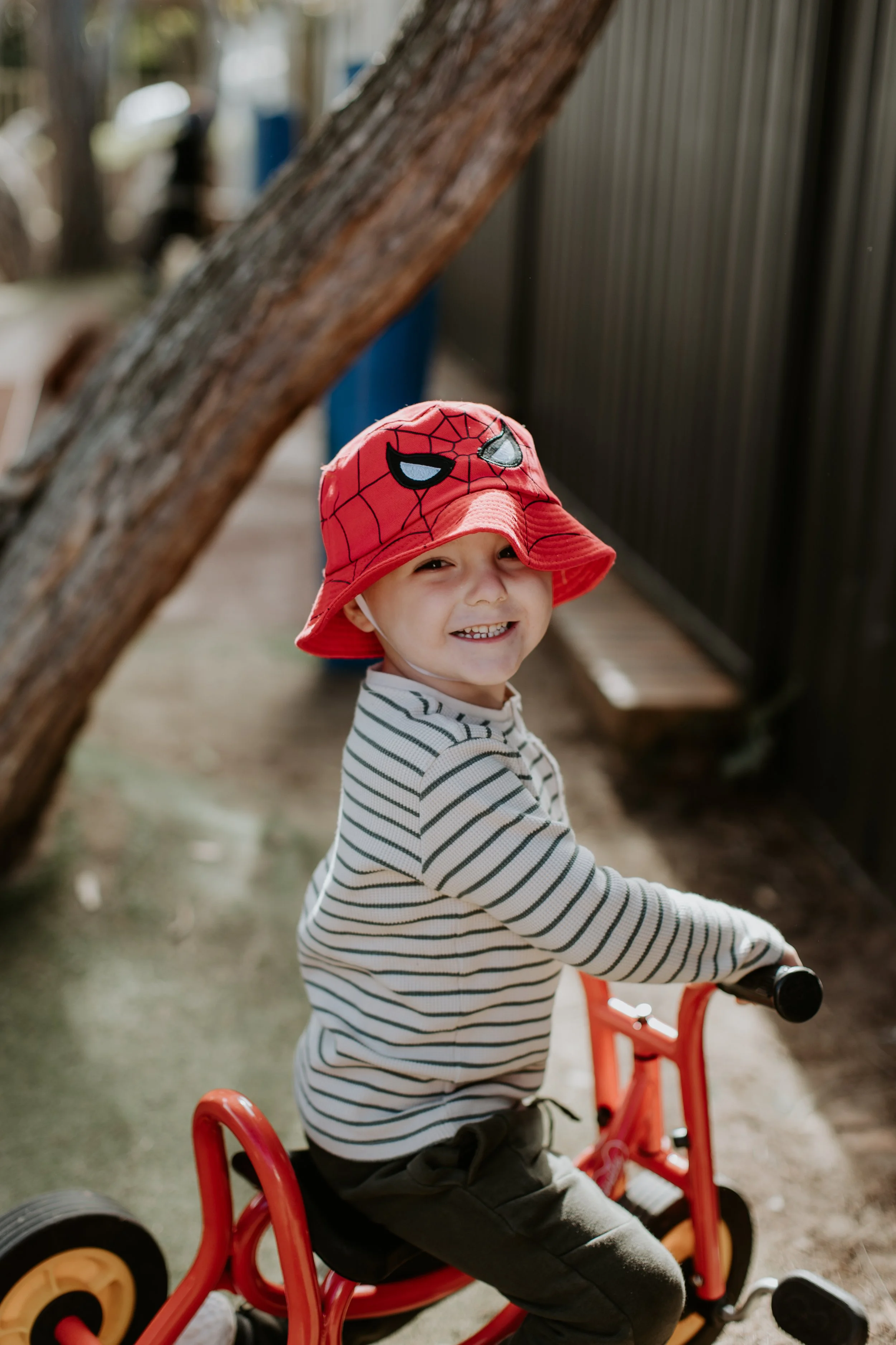 A young boy wearing a red Spider-Man themed bucket hat, striped shirt, and black pants, riding a small tricycle outdoors near a tree and a black fence.