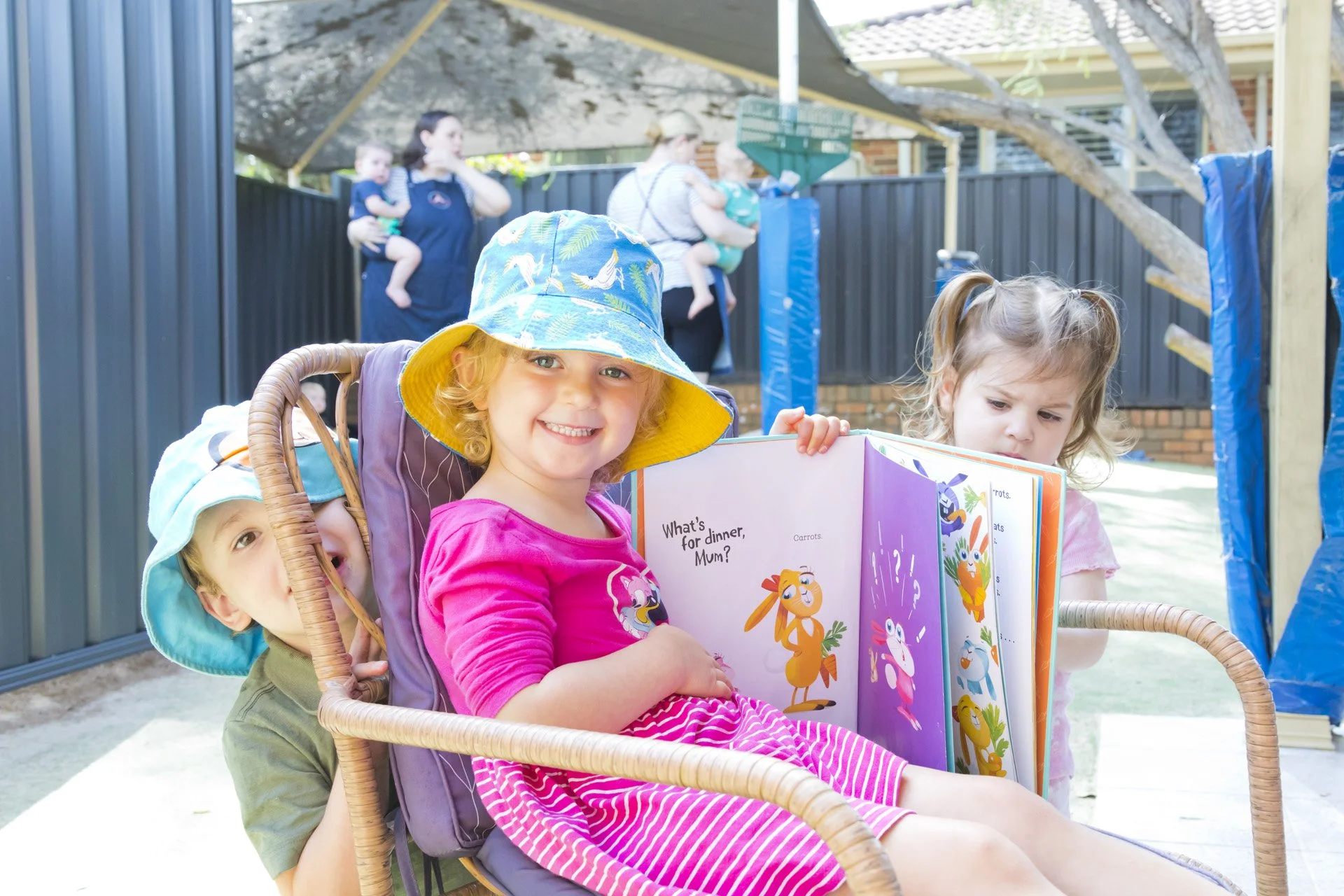 Three children sitting on a porch, one girl in a pink shirt reading a book, a boy behind her, and another girl next to her, with a backyard scene and adults holding children in the background.