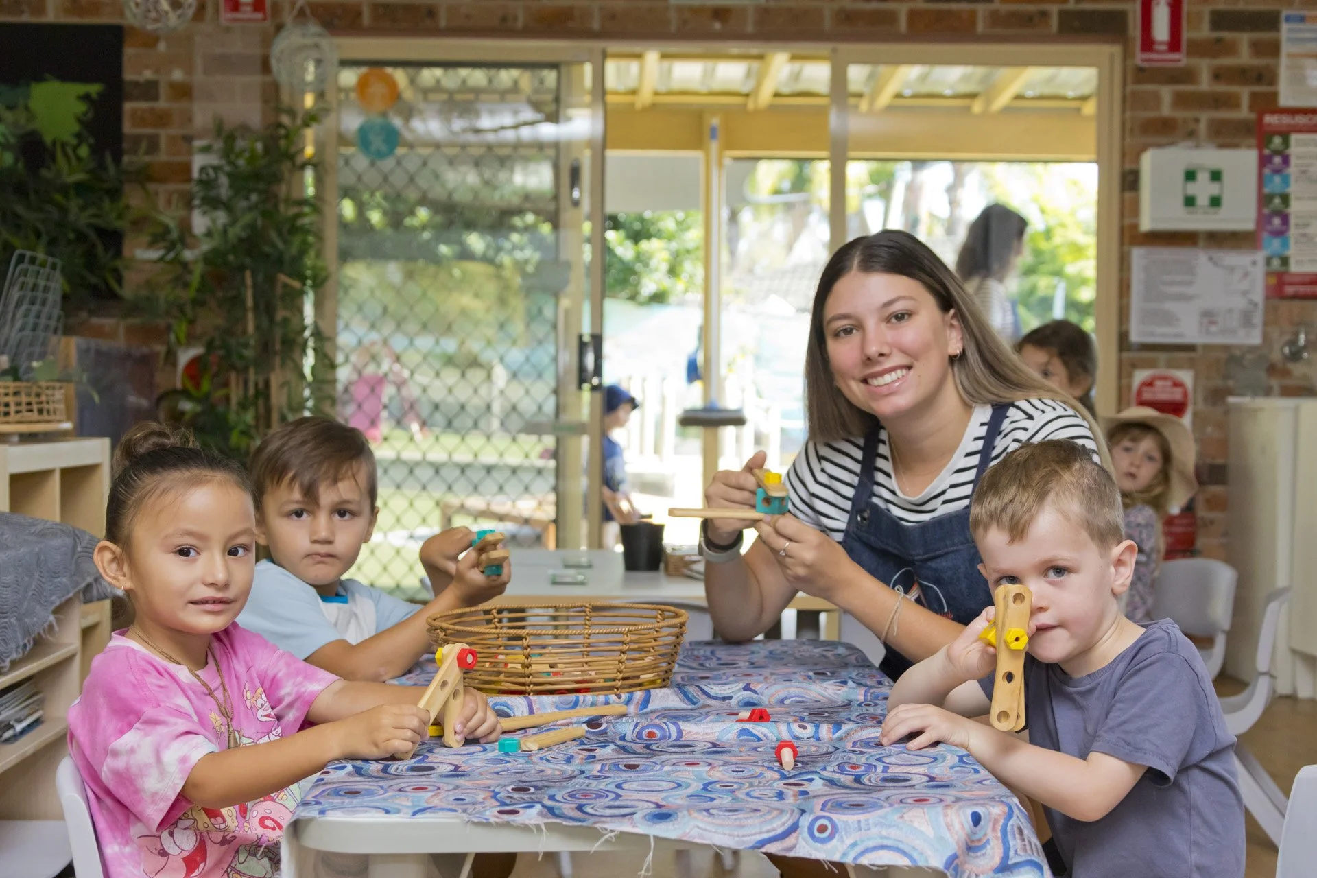 A group of young children and a woman playing with wooden toys at a table in a classroom or daycare setting, with a view of an outdoor area through a glass door.